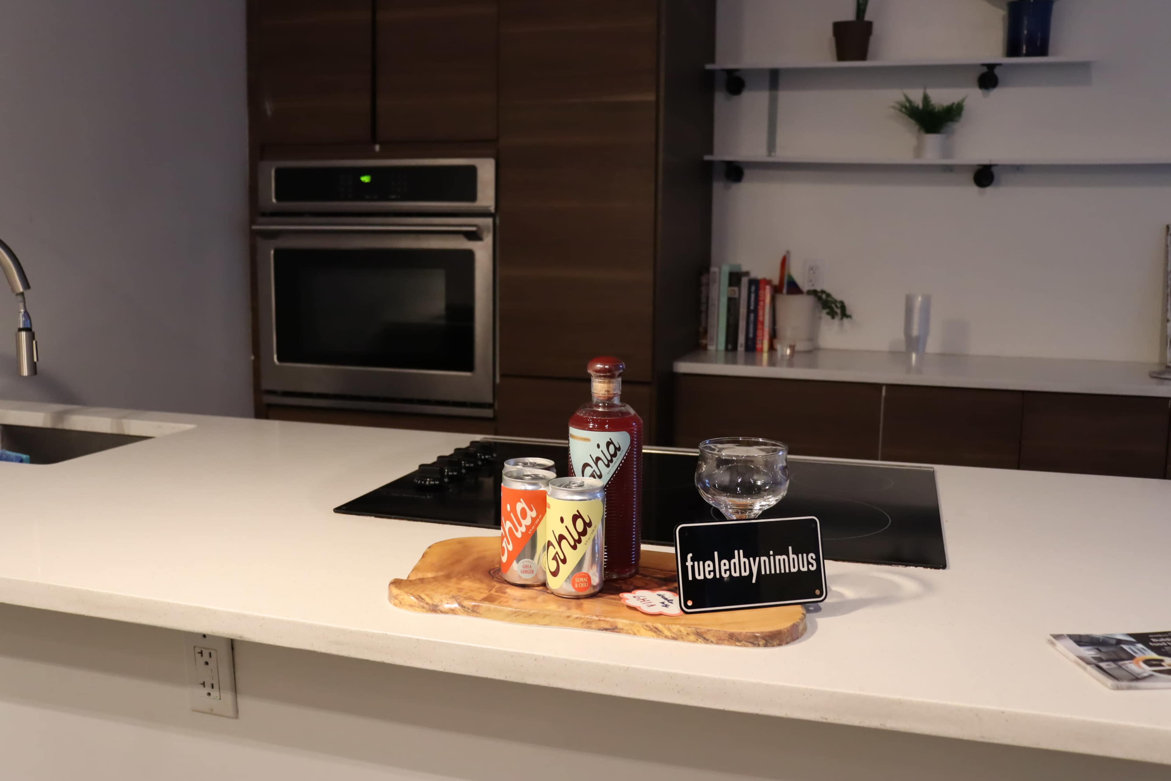A kitchen counter displays a bottle and cans of soda alongside a glass and a wooden serving board.