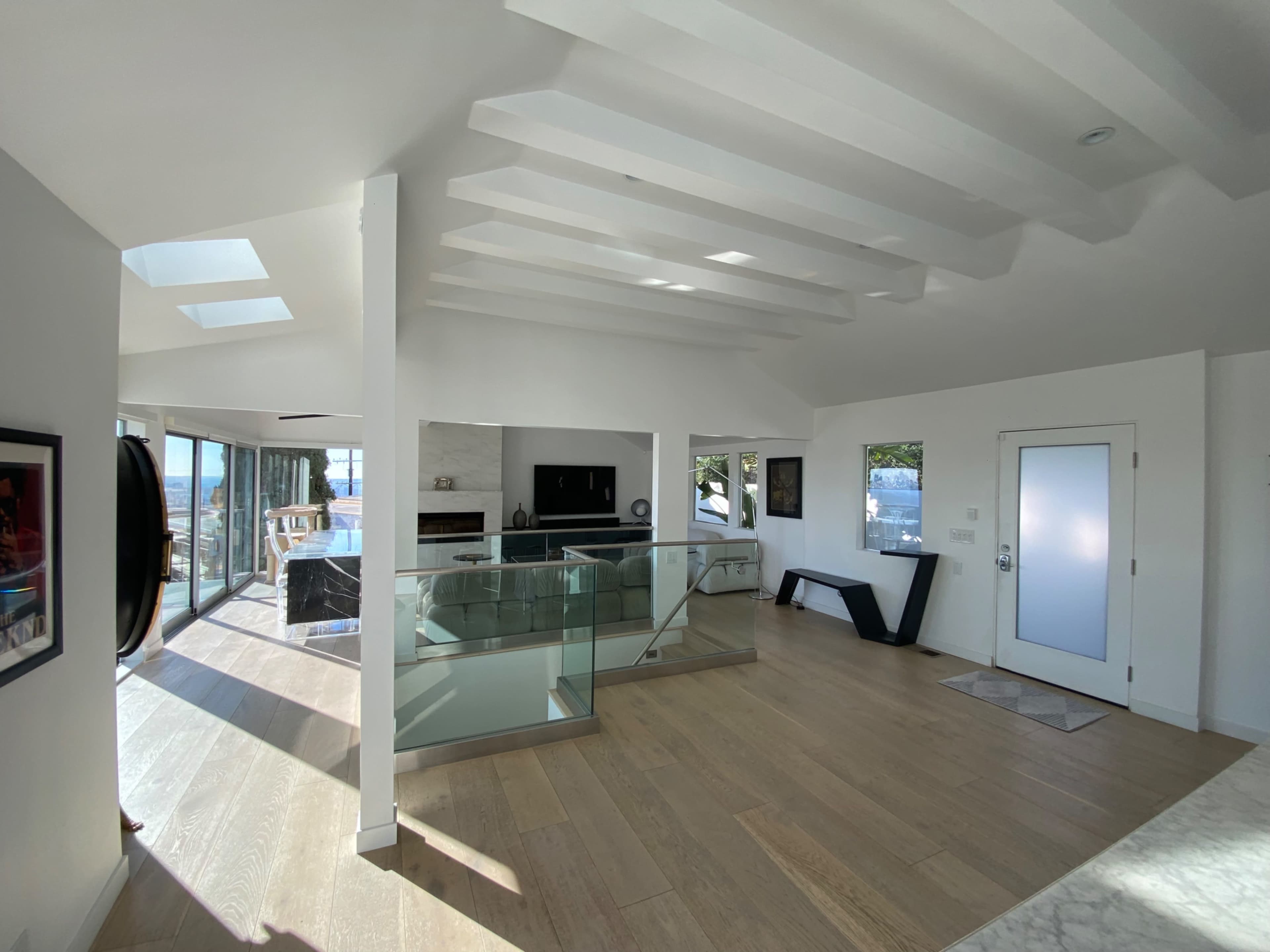 The image shows a modern interior space with an open layout, featuring wooden flooring, glass railings, and large windows that provide natural light and a view of the outdoors.