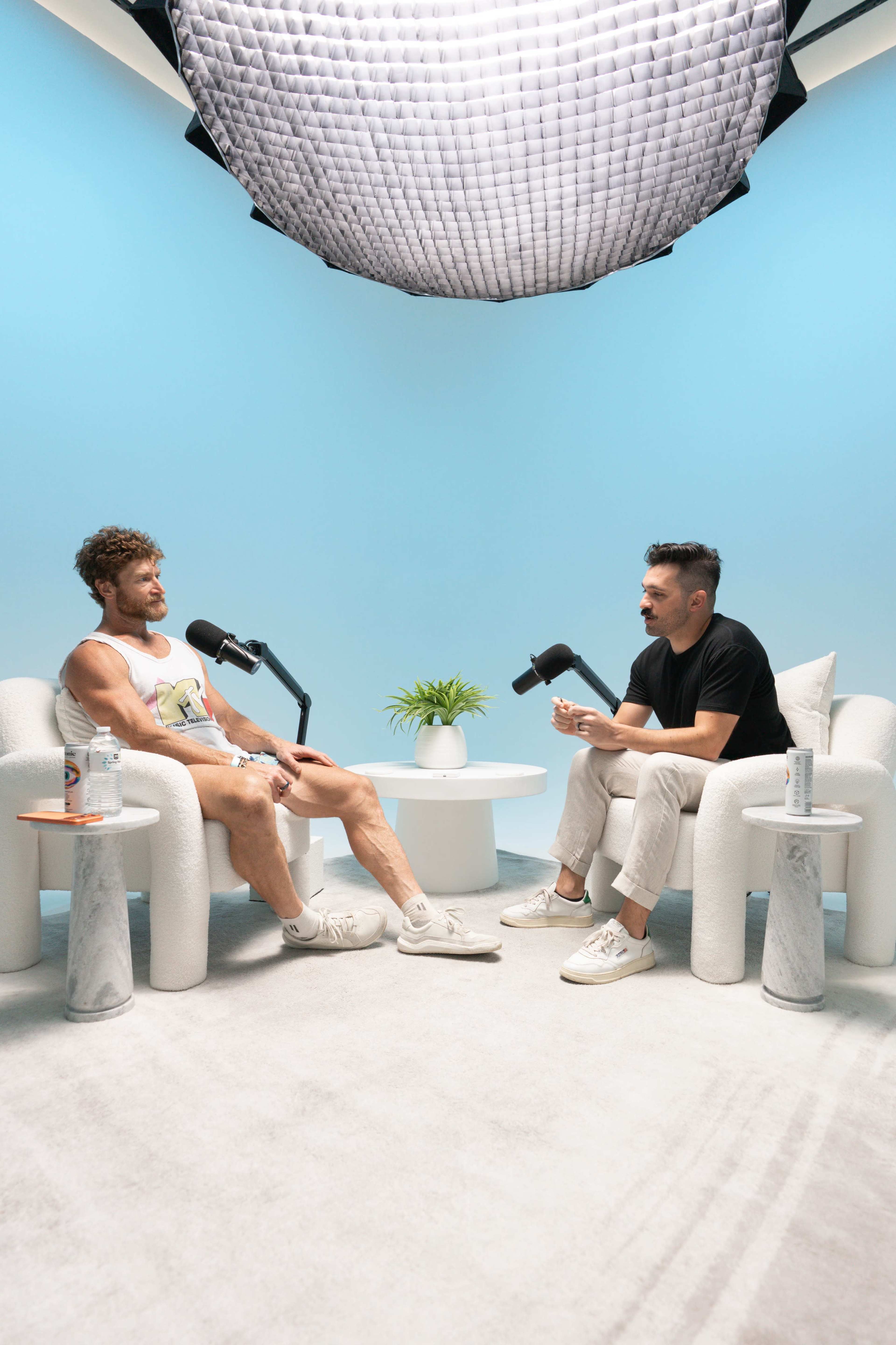 Two men are sitting in minimalist chairs with microphones in a brightly lit studio, discussing in front of a small round table with a plant and drinks.