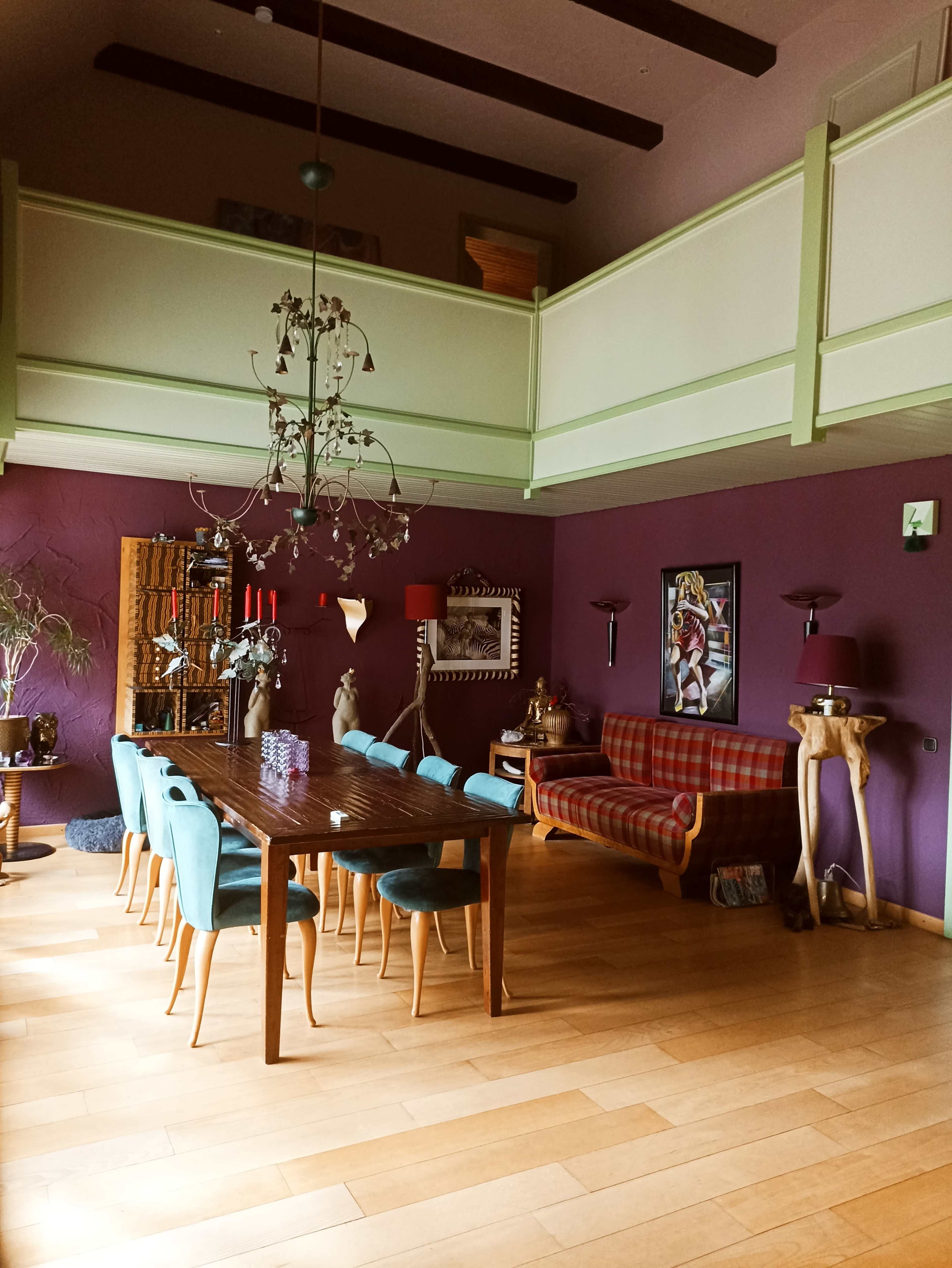 The image shows a spacious dining area with a long wooden table surrounded by turquoise chairs, a large chandelier overhead, and a cozy seating area featuring a red and yellow plaid sofa.