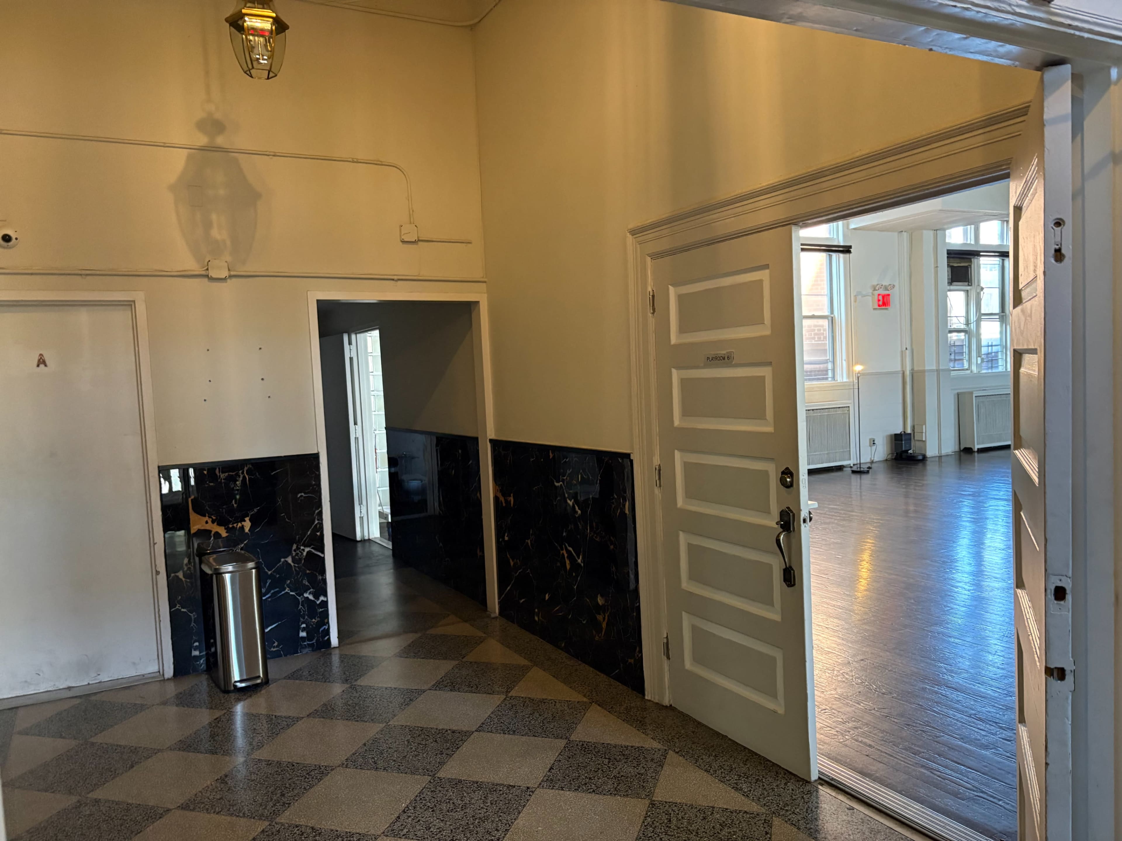 The image shows a corridor with a marbled wall leading to an open door that opens into a well-lit room with wooden floors.