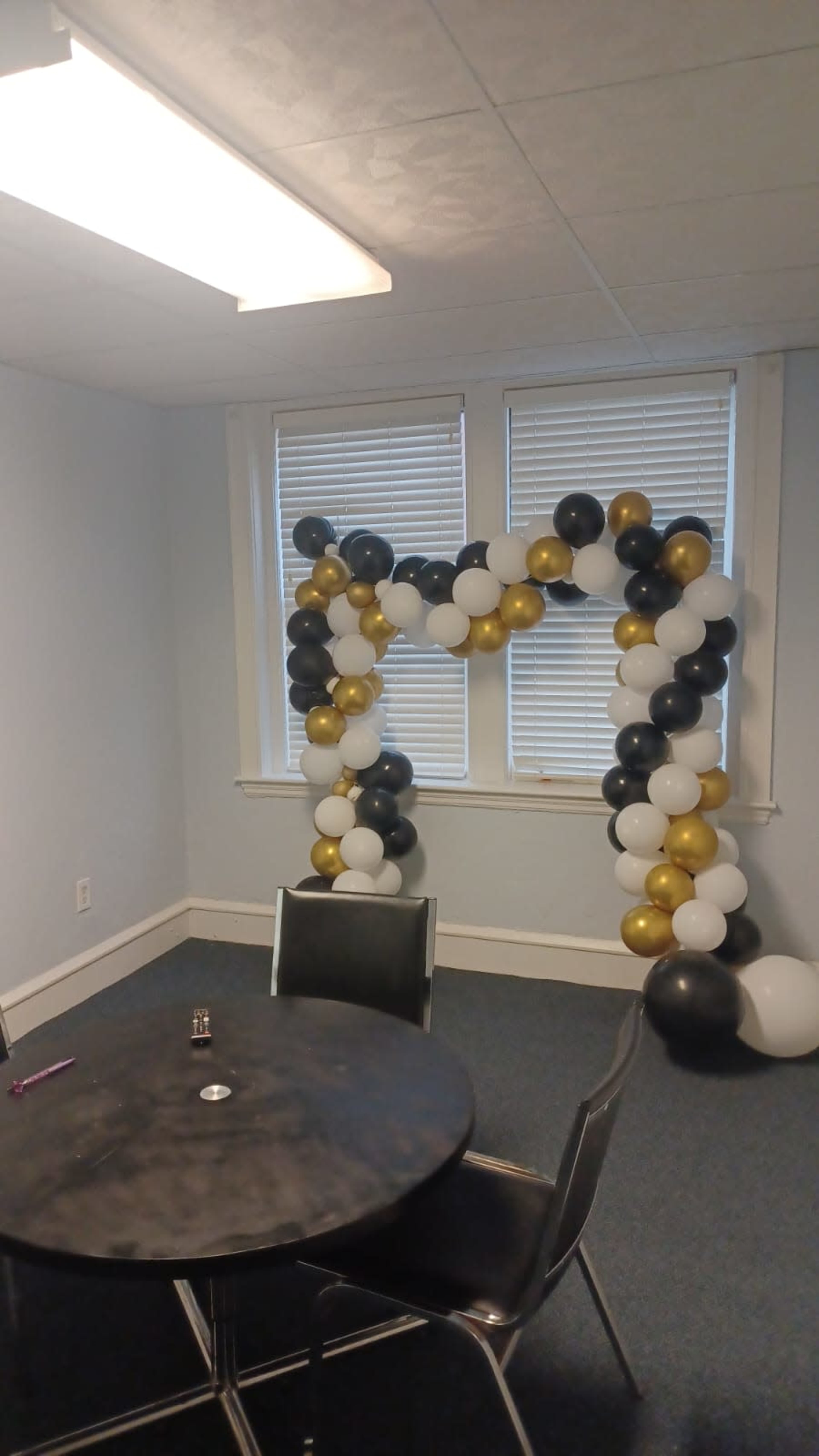 A balloon arch in black, gold, and white decorates a corner of a room with a table and chairs.
