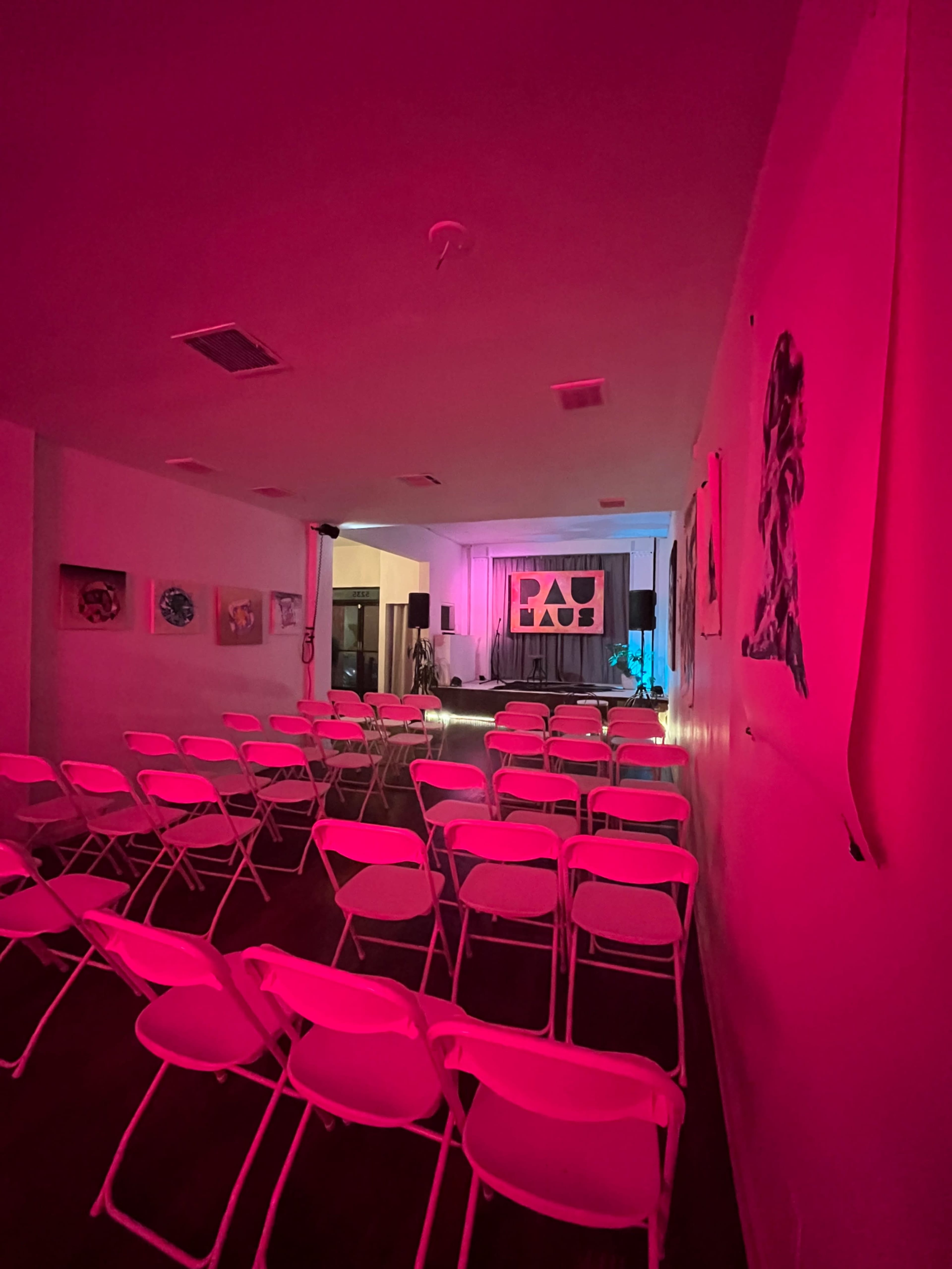 A brightly lit event space features neatly arranged white folding chairs facing a stage with a backdrop and artwork on the walls.