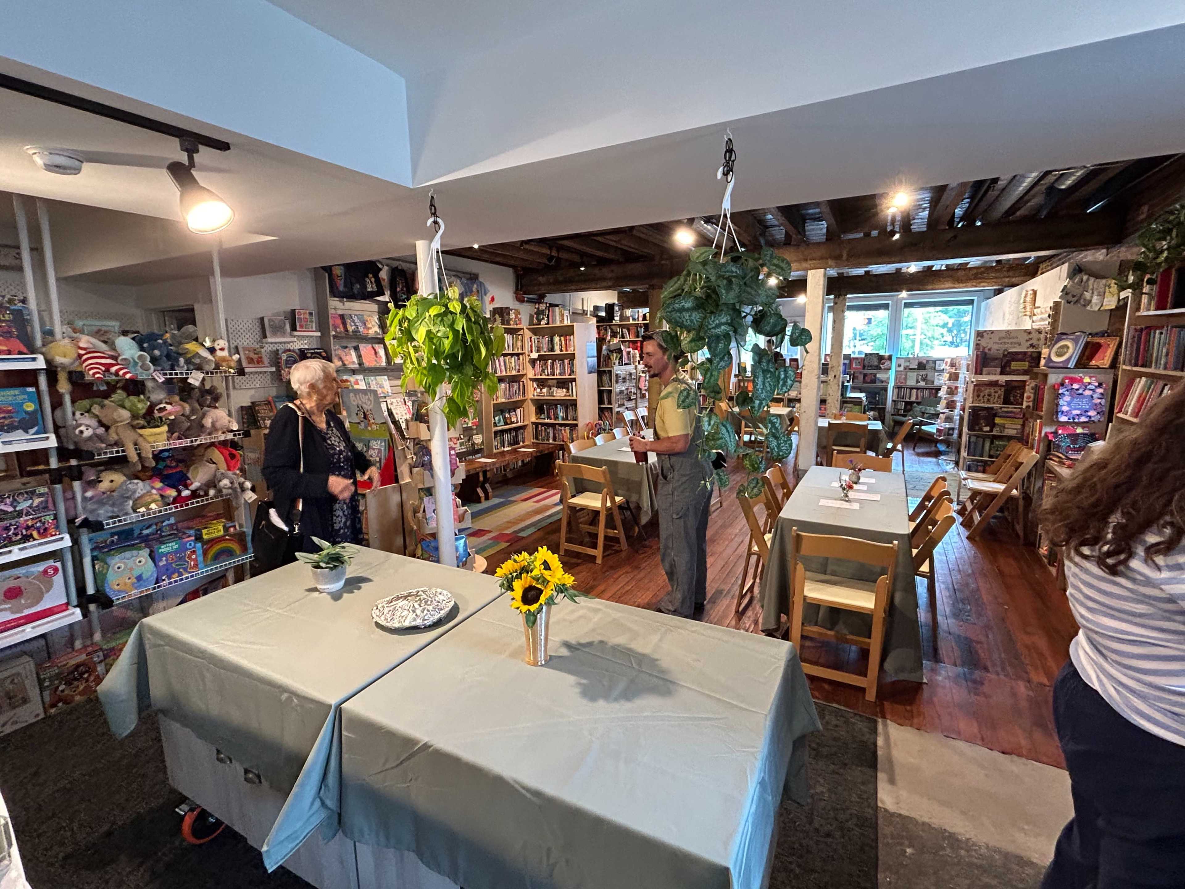 The image shows a cozy bookstore with wooden beams, shelves filled with books, and tables arranged for a gathering, featuring potted plants and a few people interacting.