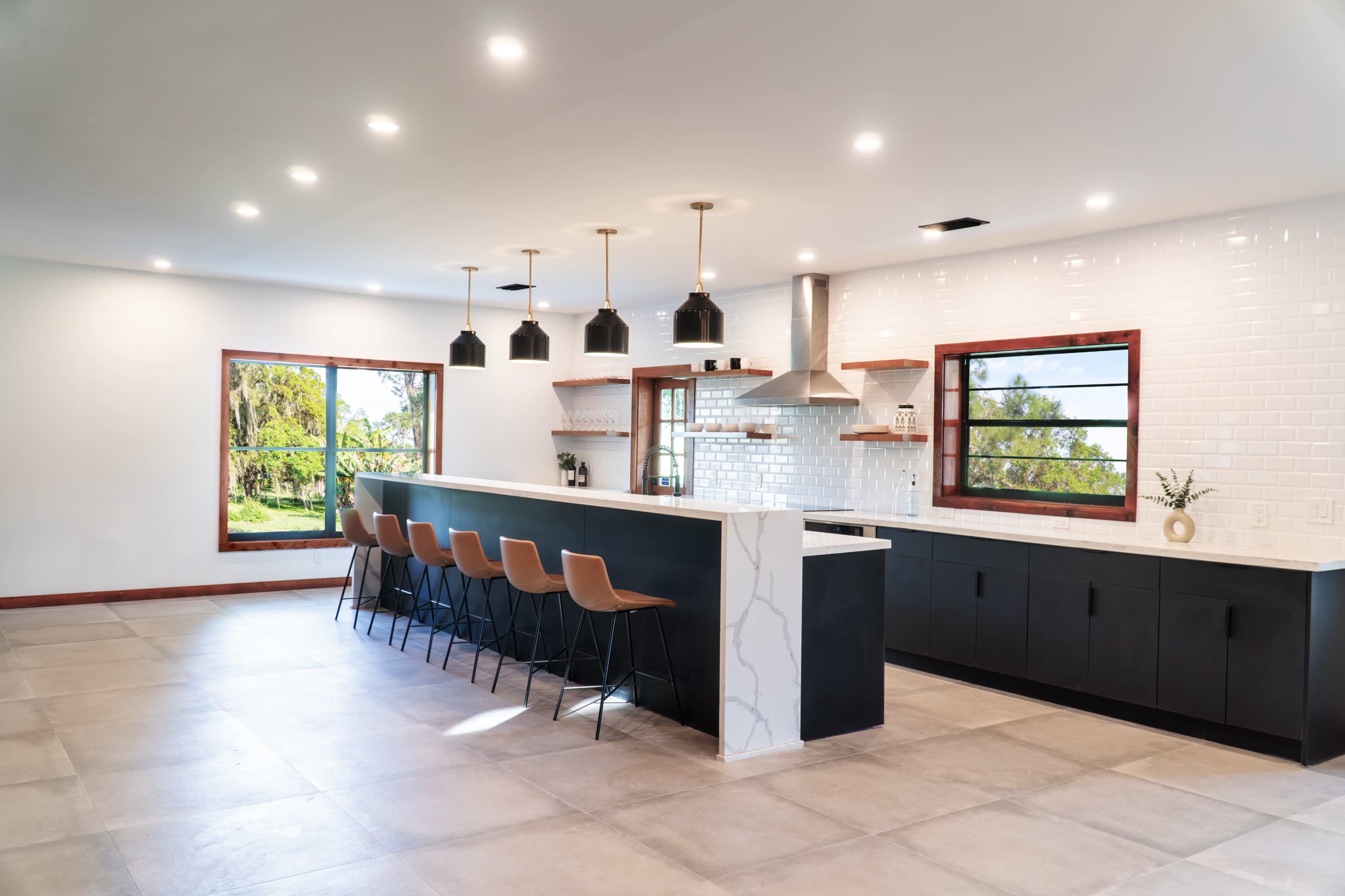 A modern kitchen features a large island with bar seating, pendant lights, and windows that overlook a landscaped area.
