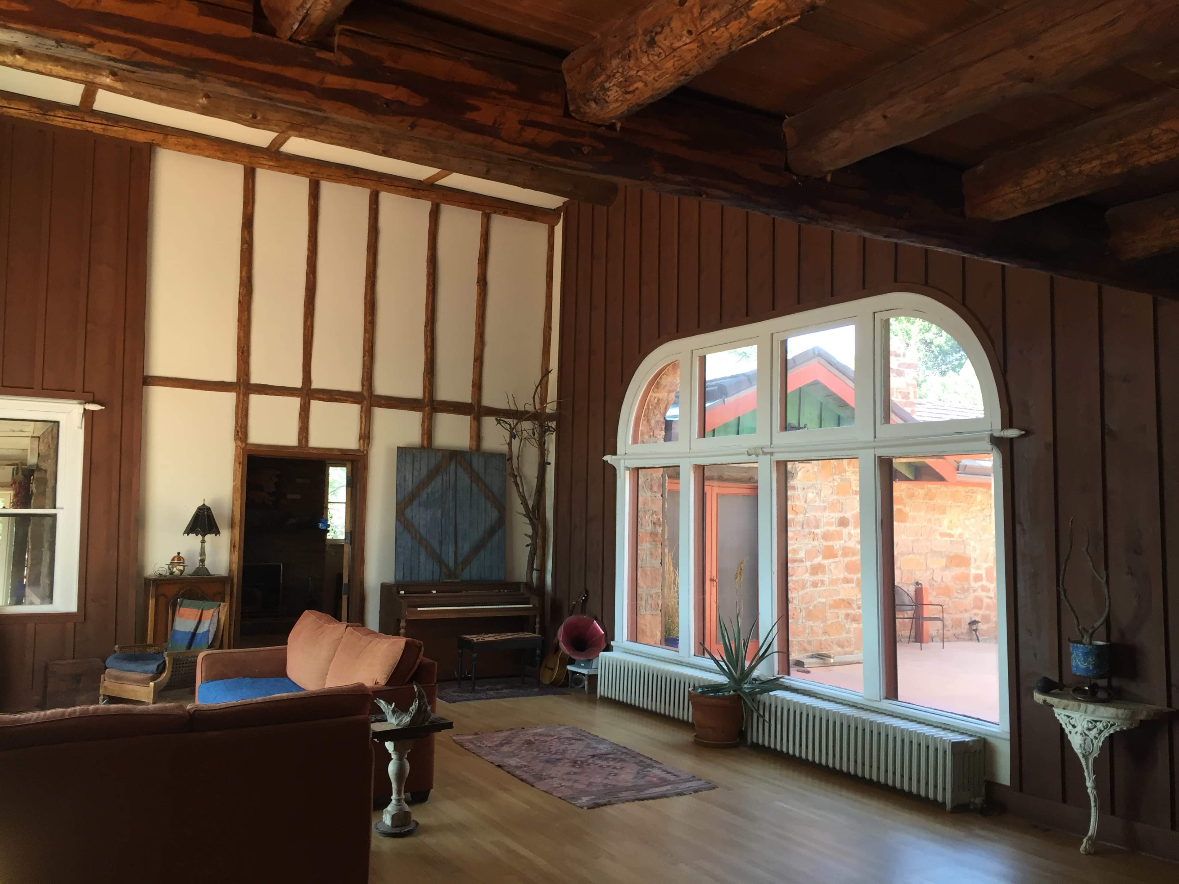 A spacious living room with wooden beams, a large arched window, a piano, and a plants placed on a radiator and a small table.