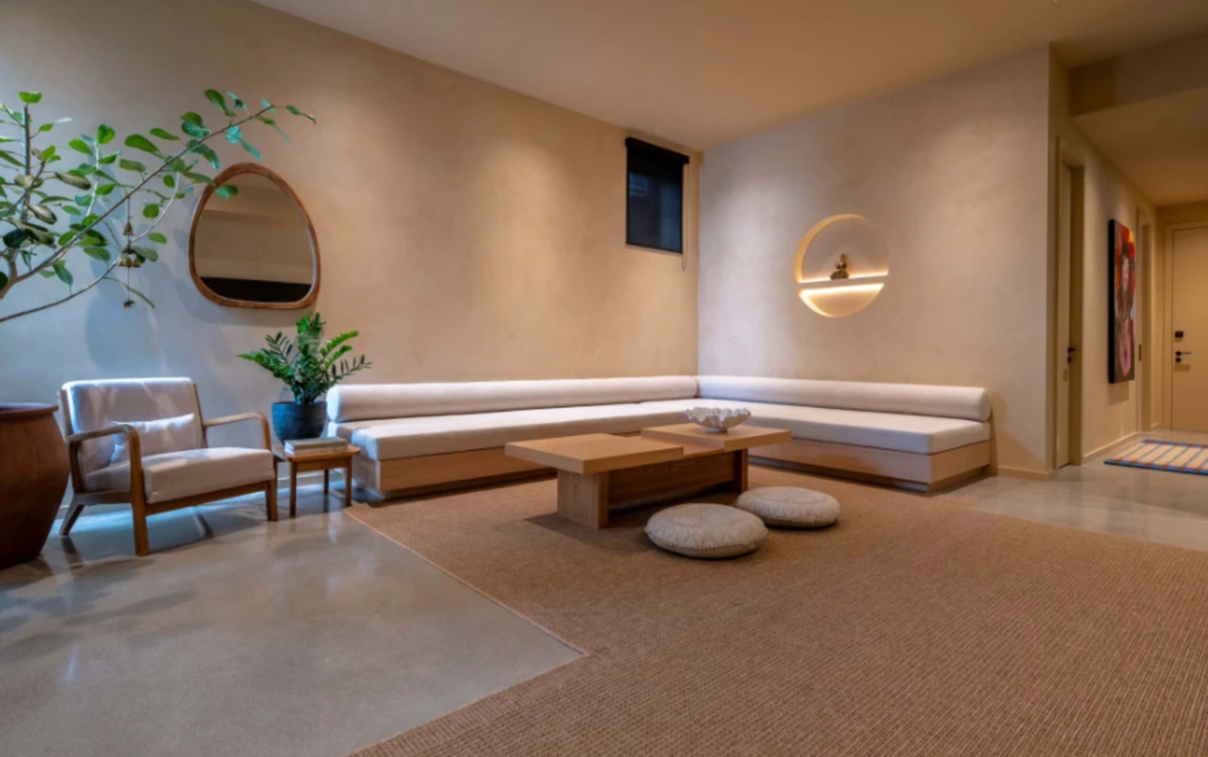 The image shows a minimalist living area featuring a low wood table, a sectional couch, a potted plant, and simple seating on a woven rug.