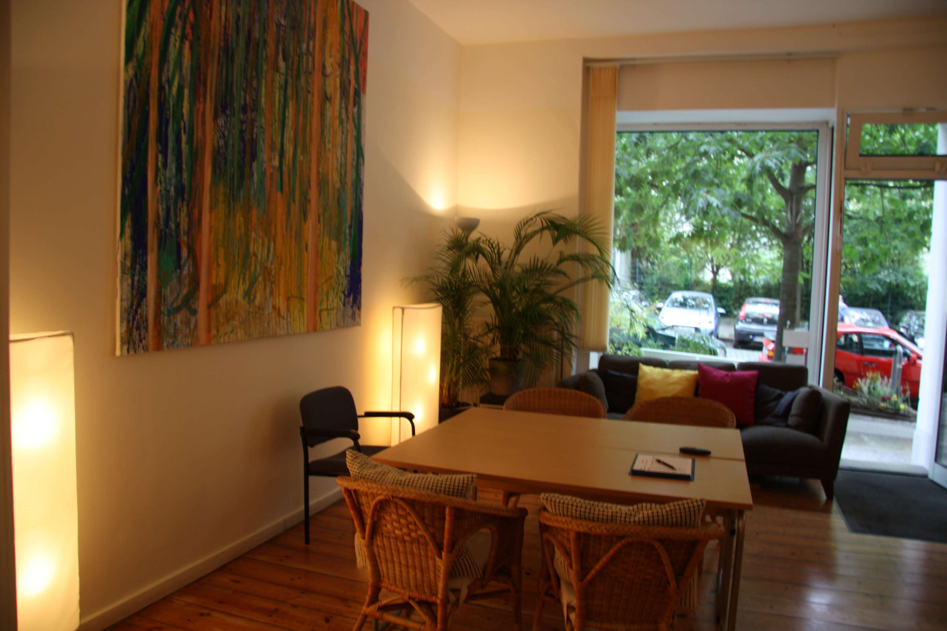 A bright room features a large abstract painting on one wall, a wooden table surrounded by chairs, and a view of greenery and parked cars through a large window.