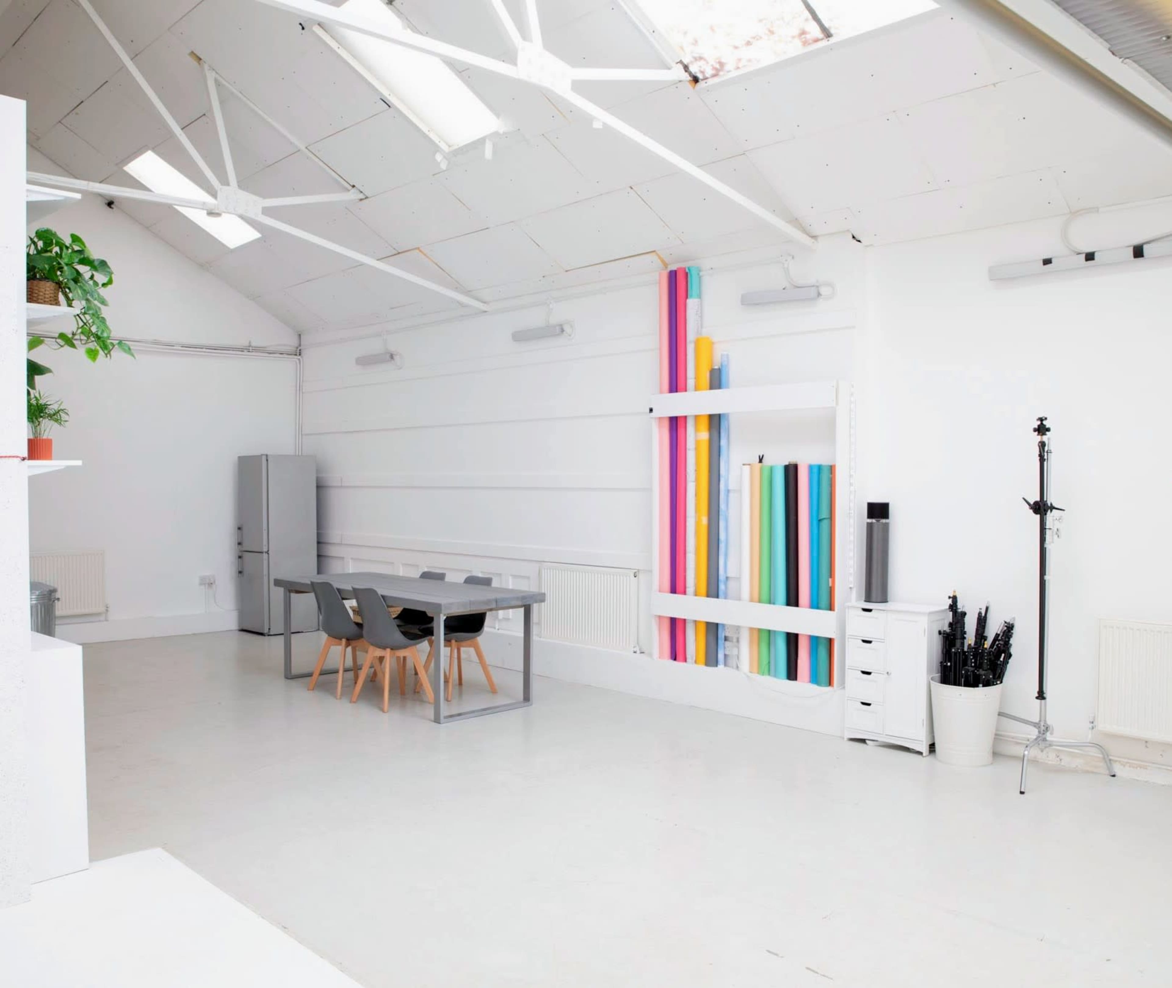 The image shows a bright, minimalist studio space with a large table and colorful rolls of paper displayed against a wall.