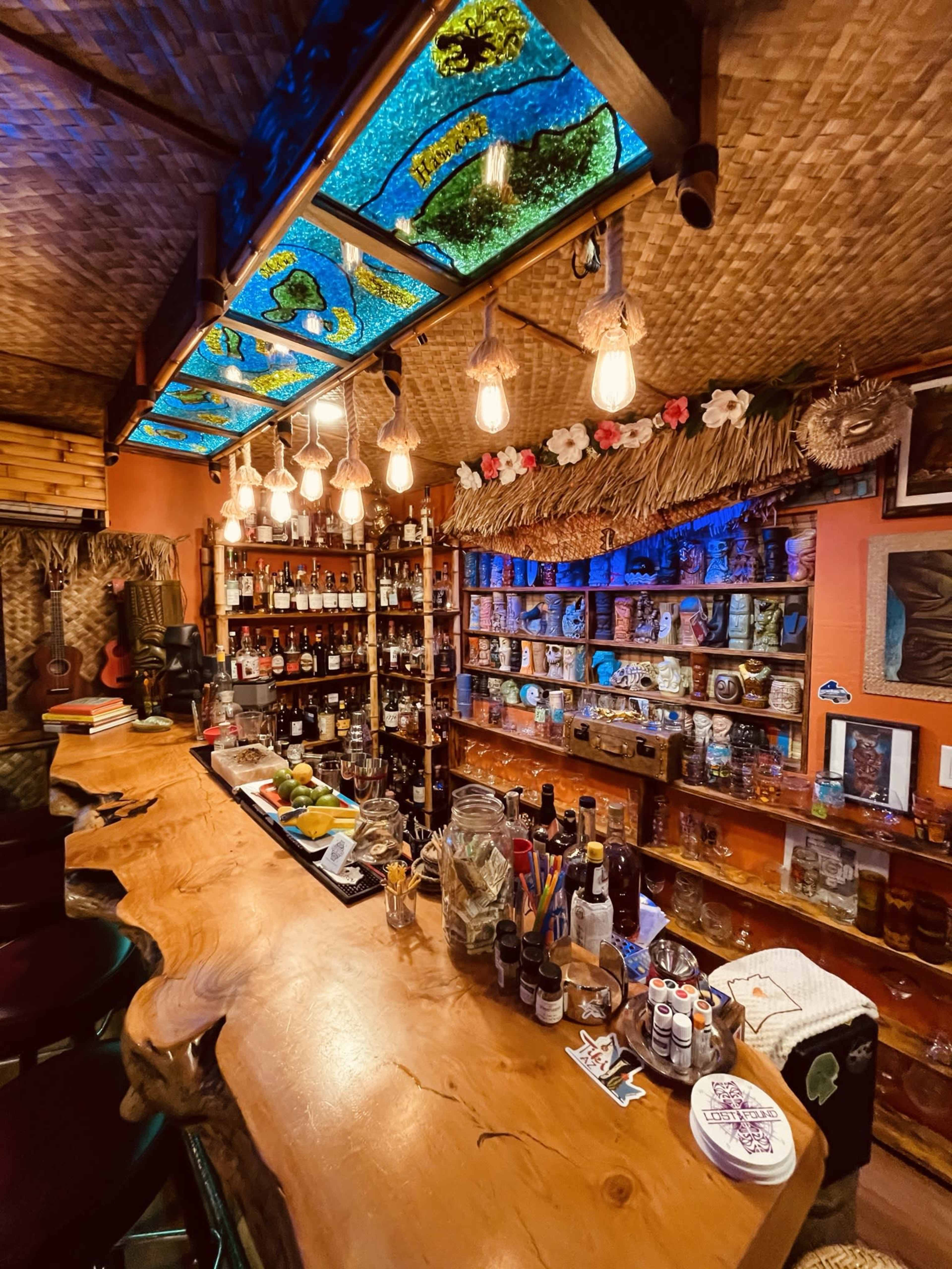 A well-decorated bar features a long wooden countertop, shelves filled with various bottles, and colorful stained glass overhead lighting.