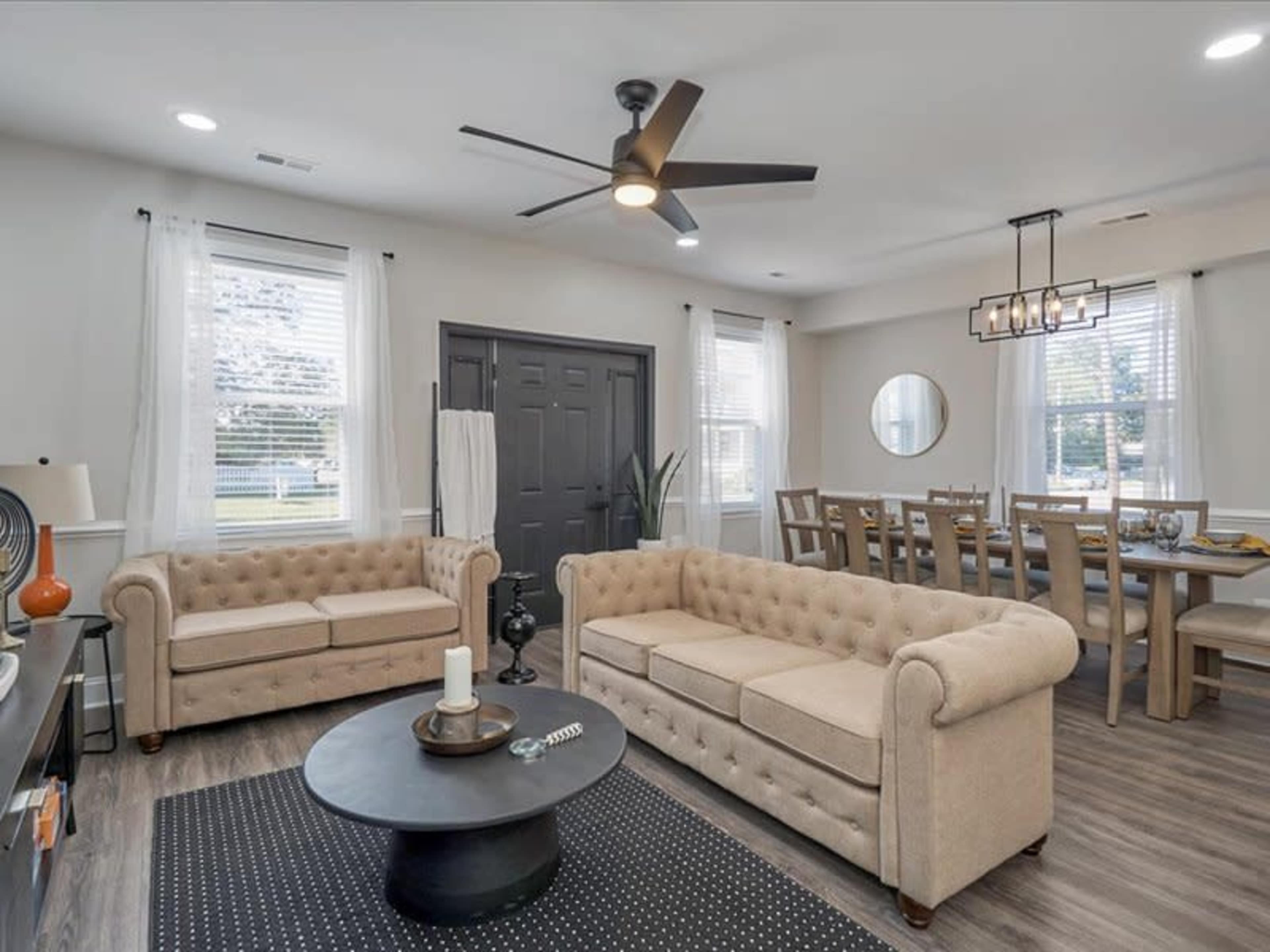 The room features two tufted beige sofas, a circular coffee table, and a dining area with a long wooden table and light fixtures.