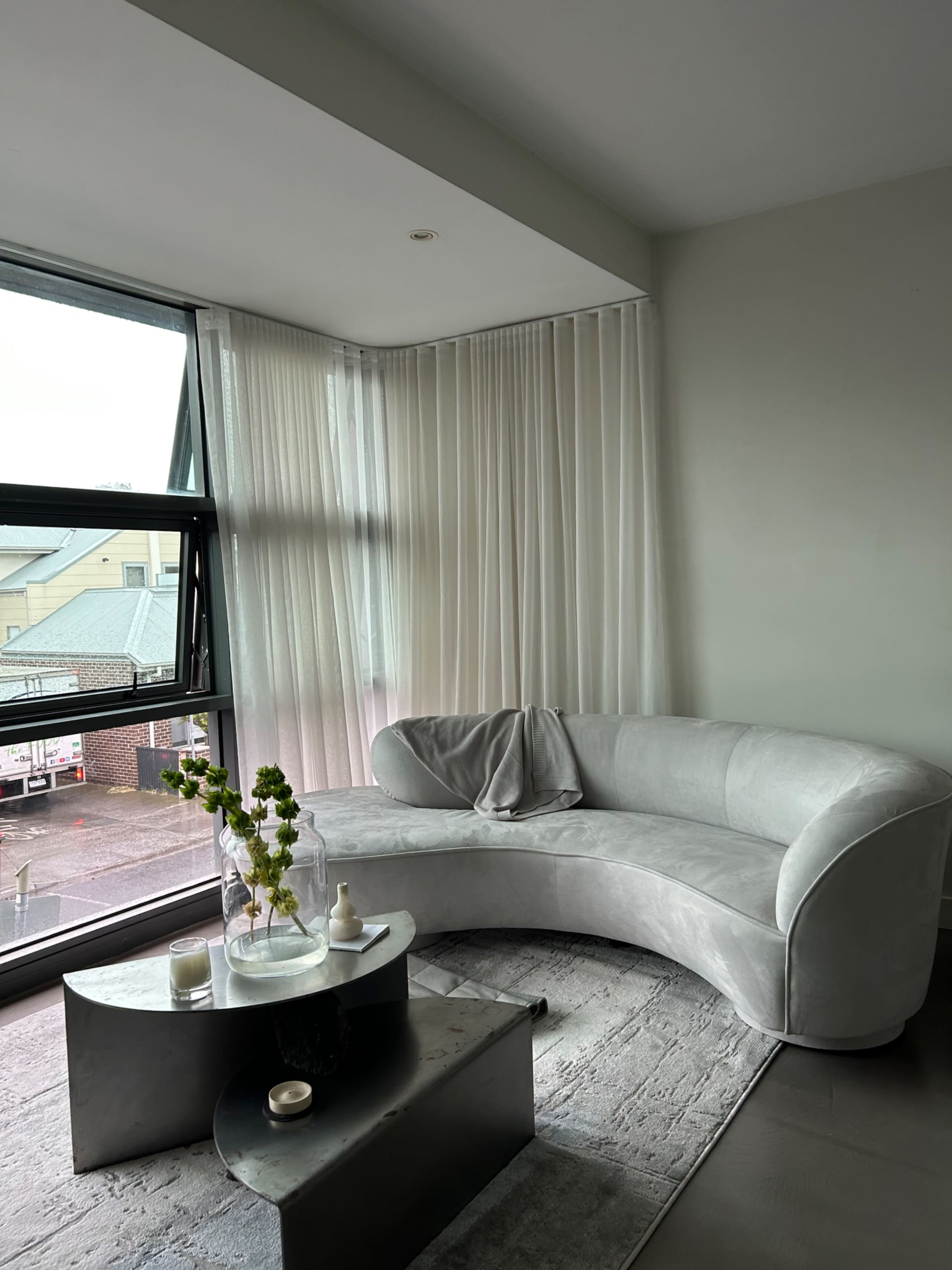 A curved gray sofa is positioned next to a large window with sheer curtains, accompanied by a marble coffee table and decorative elements in a modern living space.