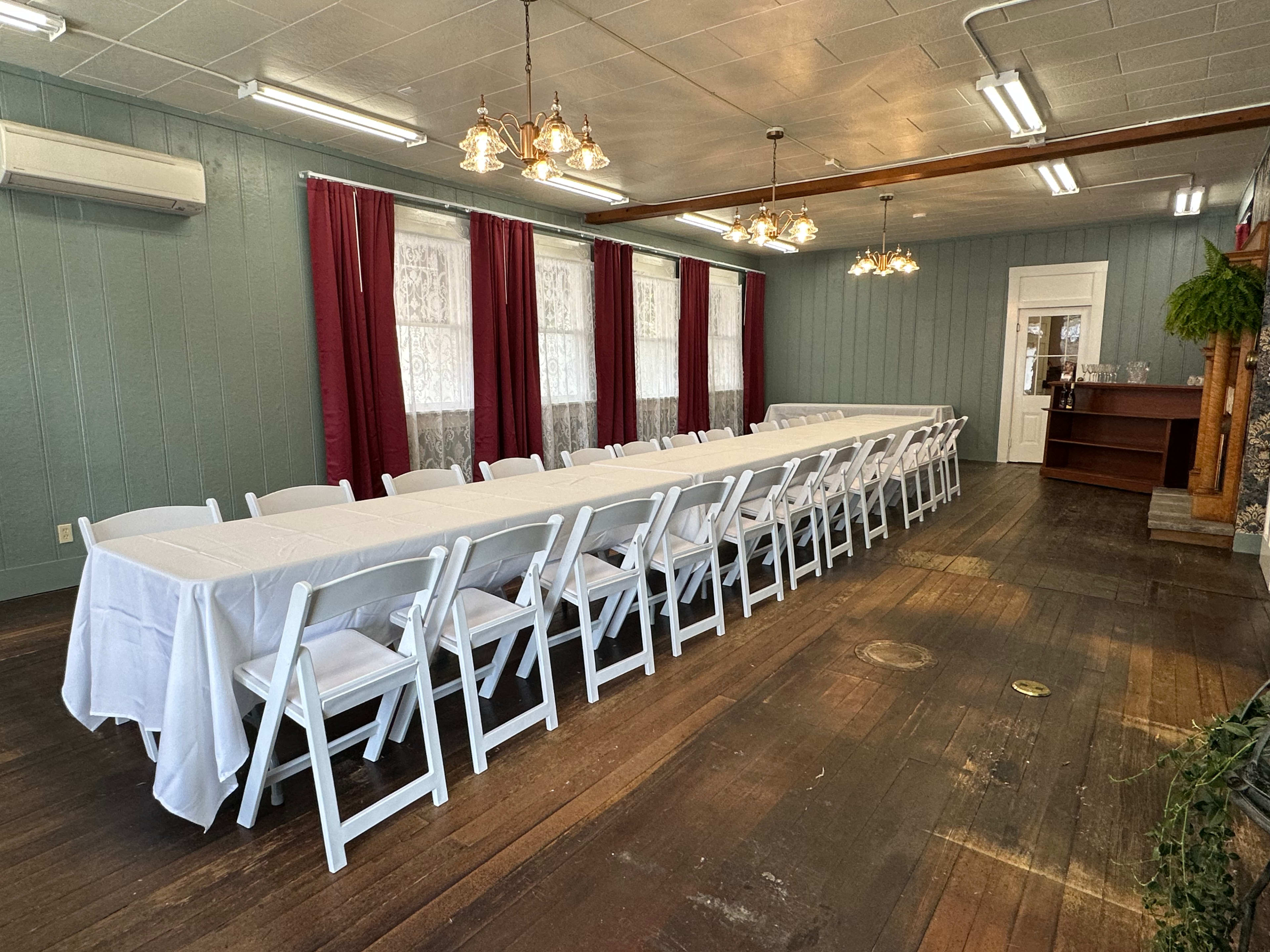 The image shows a long rectangular table covered with a white tablecloth, set with white chairs, in a room with light blue walls and large windows with red curtains.