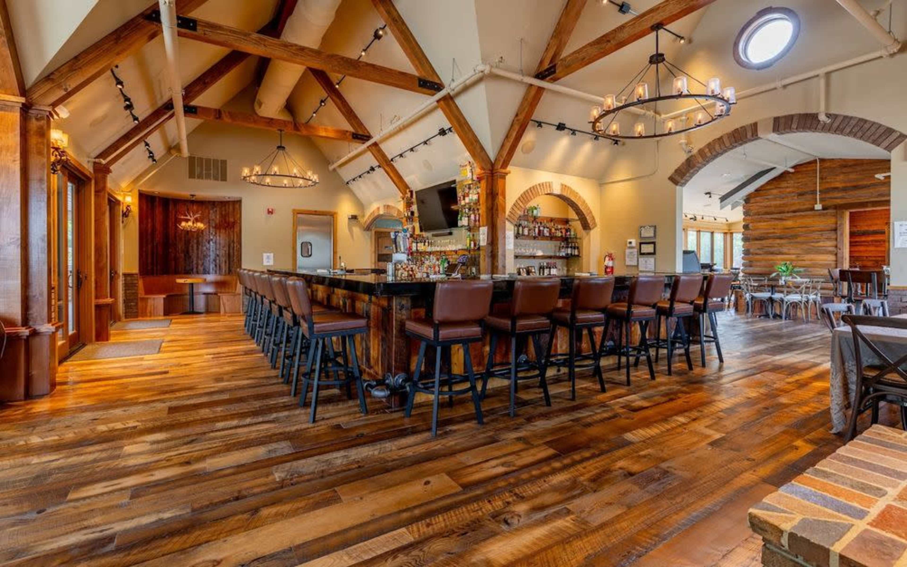 The image shows a spacious, well-lit bar area with wooden flooring, high ceilings, and a long bar counter lined with high-backed chairs.
