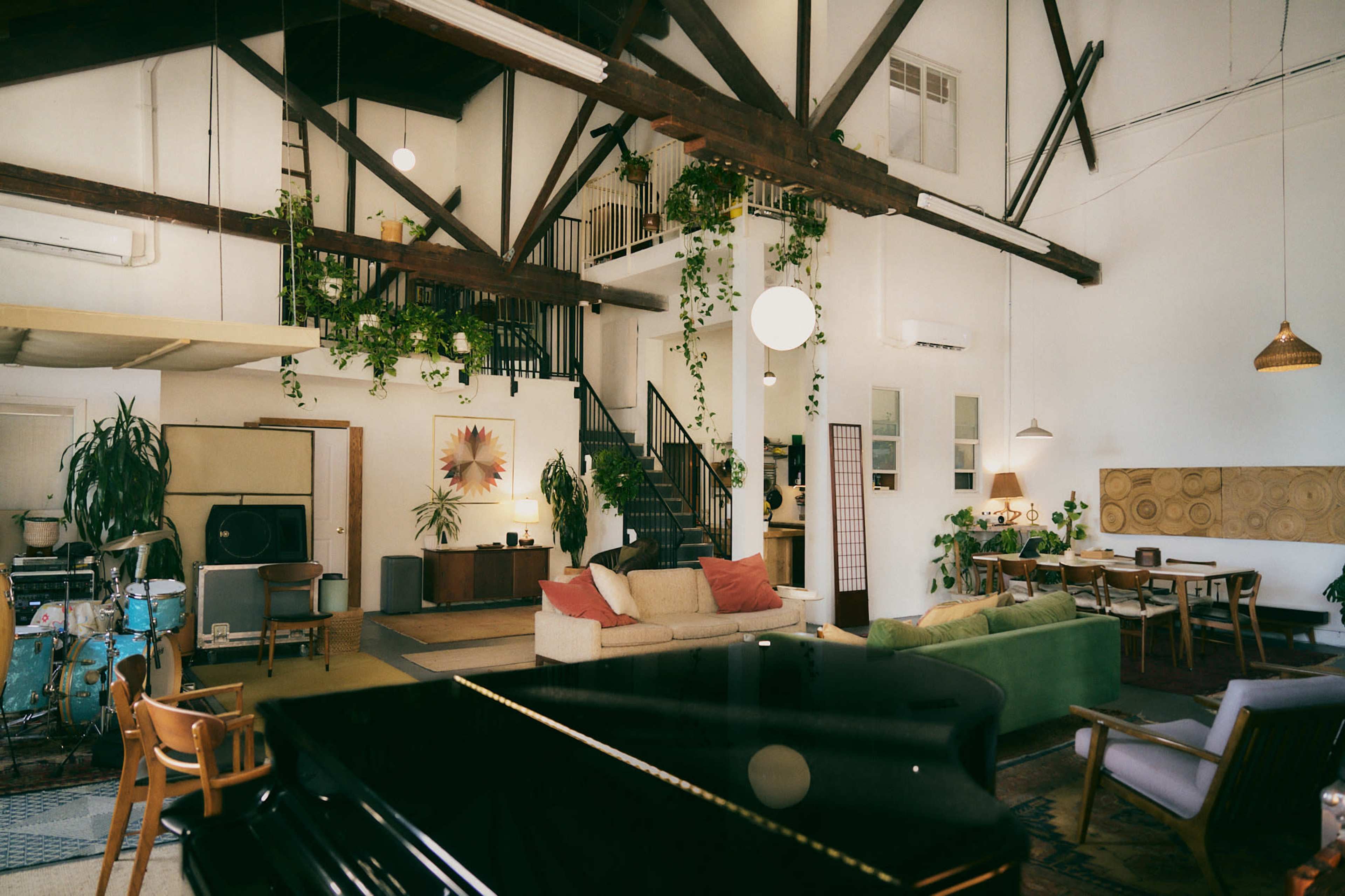 The image shows a spacious, airy living area with high ceilings, large windows, a mix of modern and vintage furniture, and various houseplants throughout the space.
