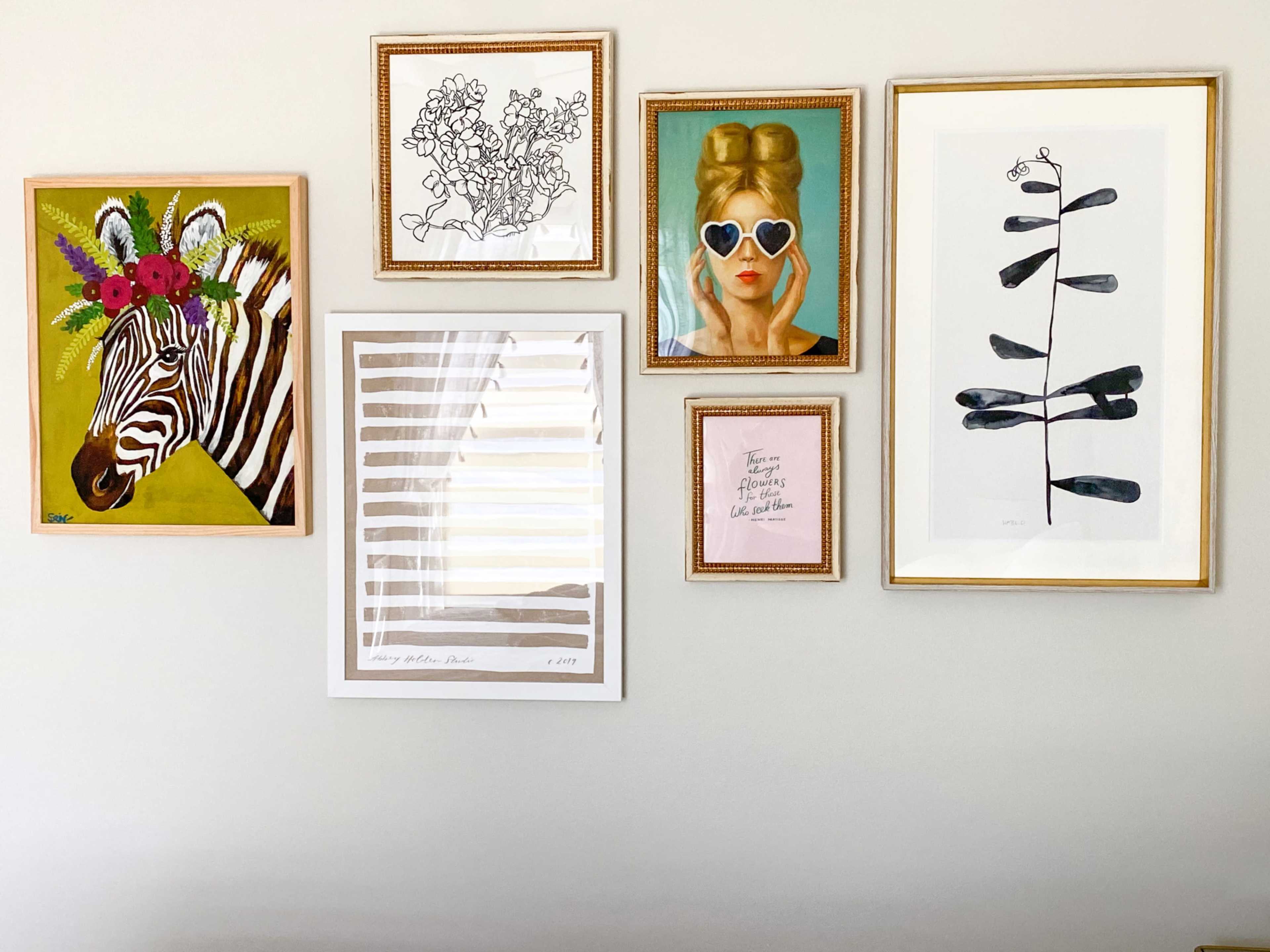 The image displays a collection of framed artwork arranged on a wall, including a zebra portrait, abstract designs, and a figure wearing oversized glasses.