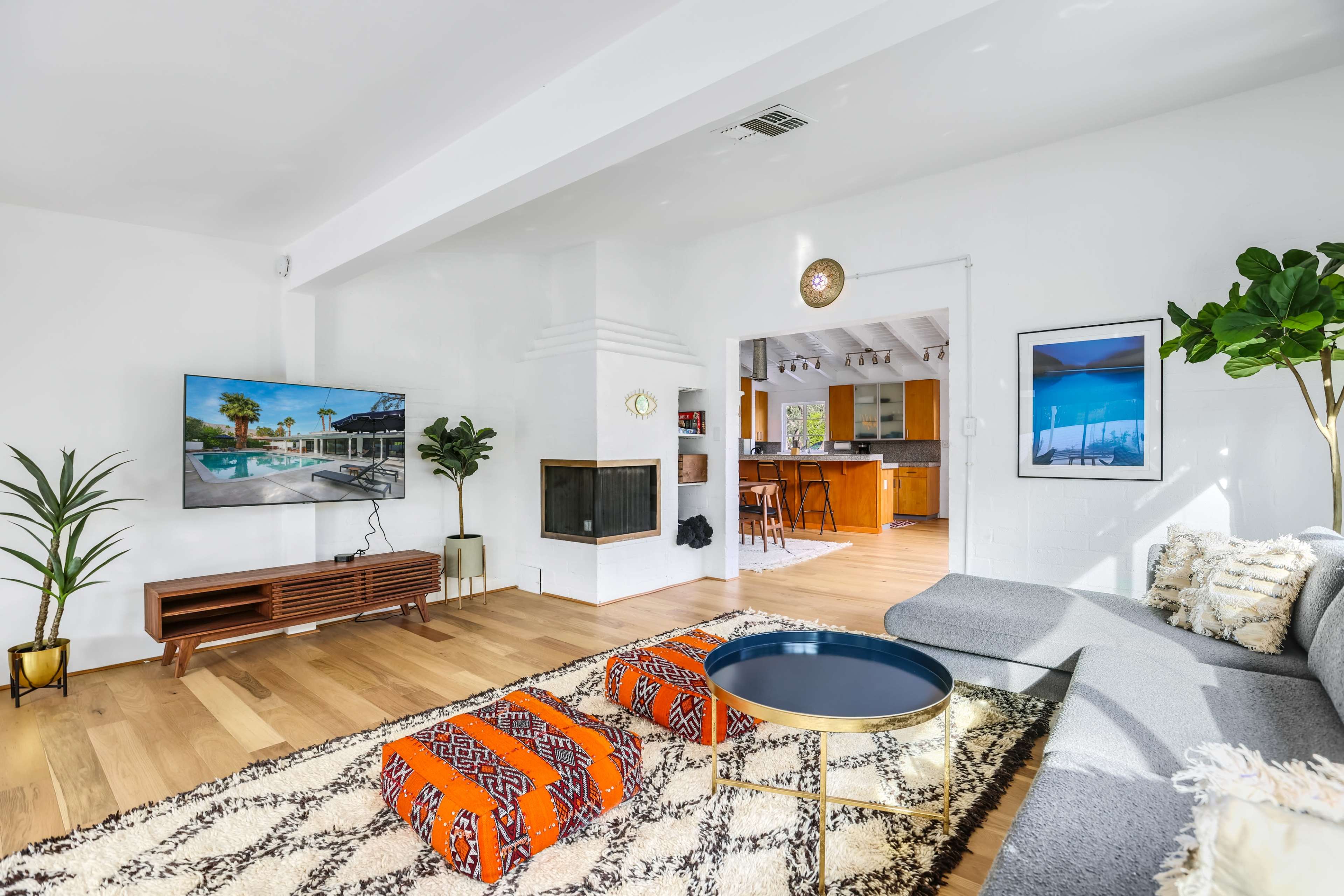 The living room features a large television mounted on the wall, a modern fireplace, a gray sofa, and a coffee table, with vibrant ottomans and plants adding decorative touches.