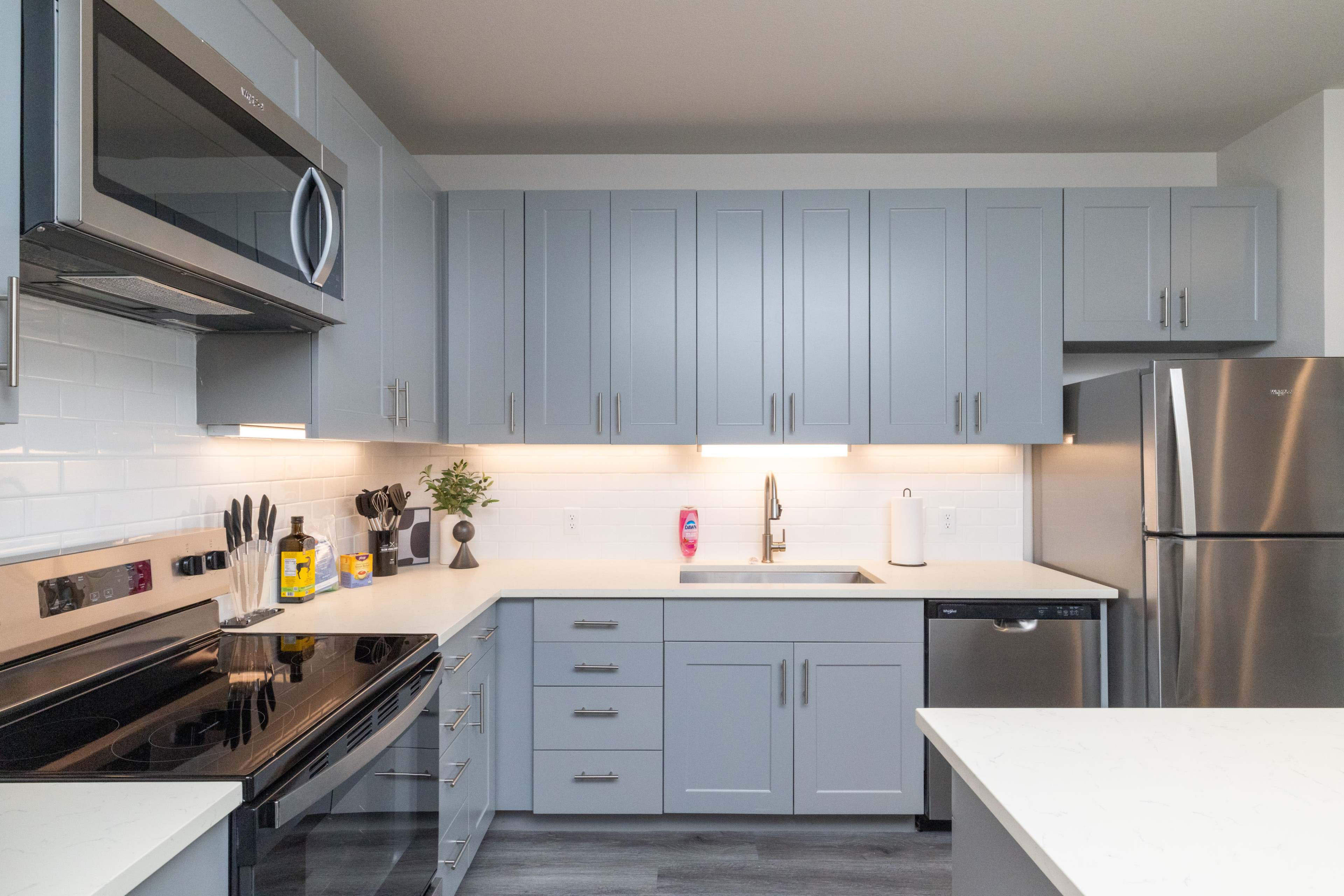 The image shows a modern kitchen with gray cabinetry, stainless steel appliances, and a white countertop.