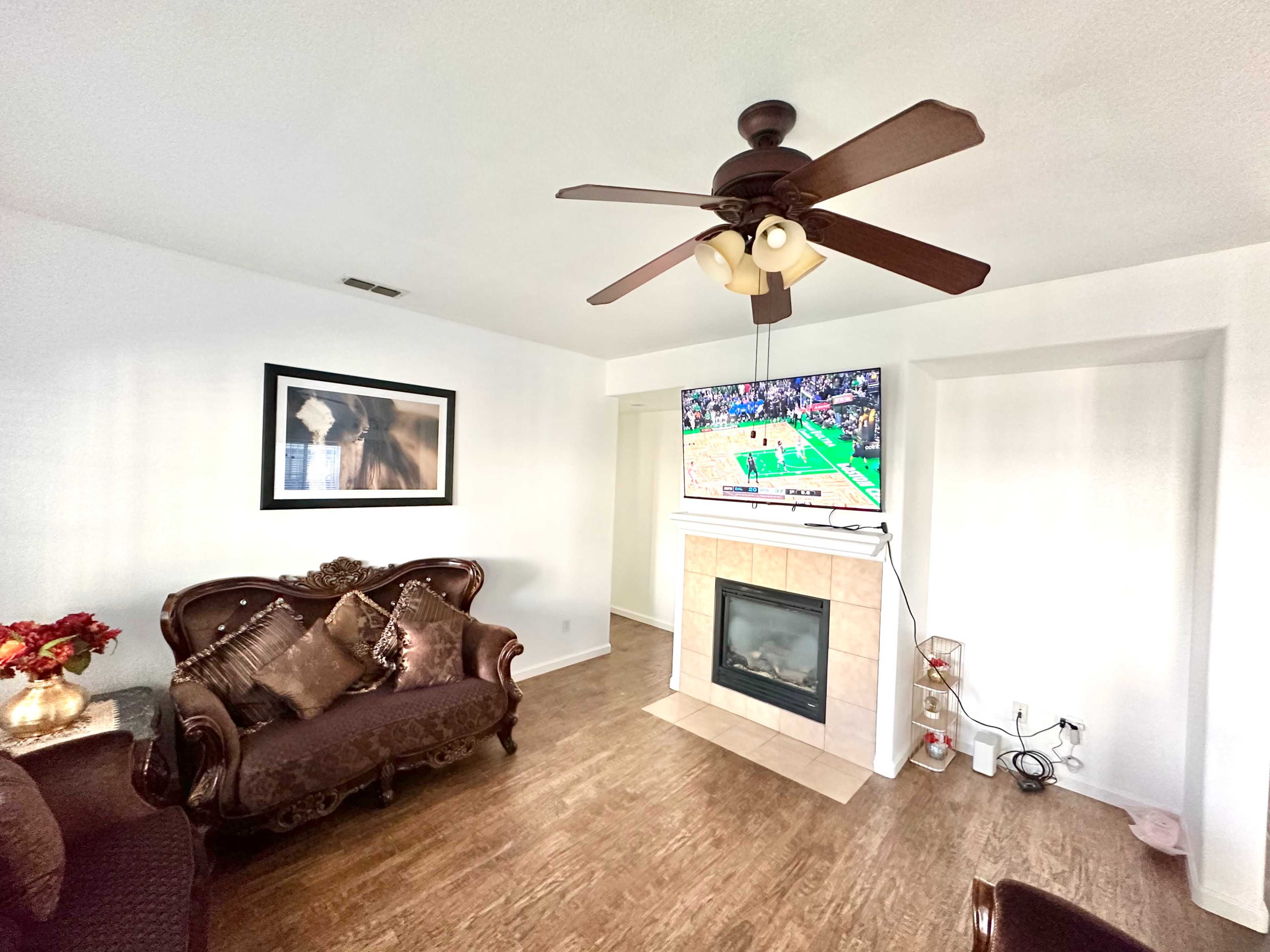 The living room features a brown couch, a mounted television displaying a basketball game, and a fireplace with decorative items nearby.