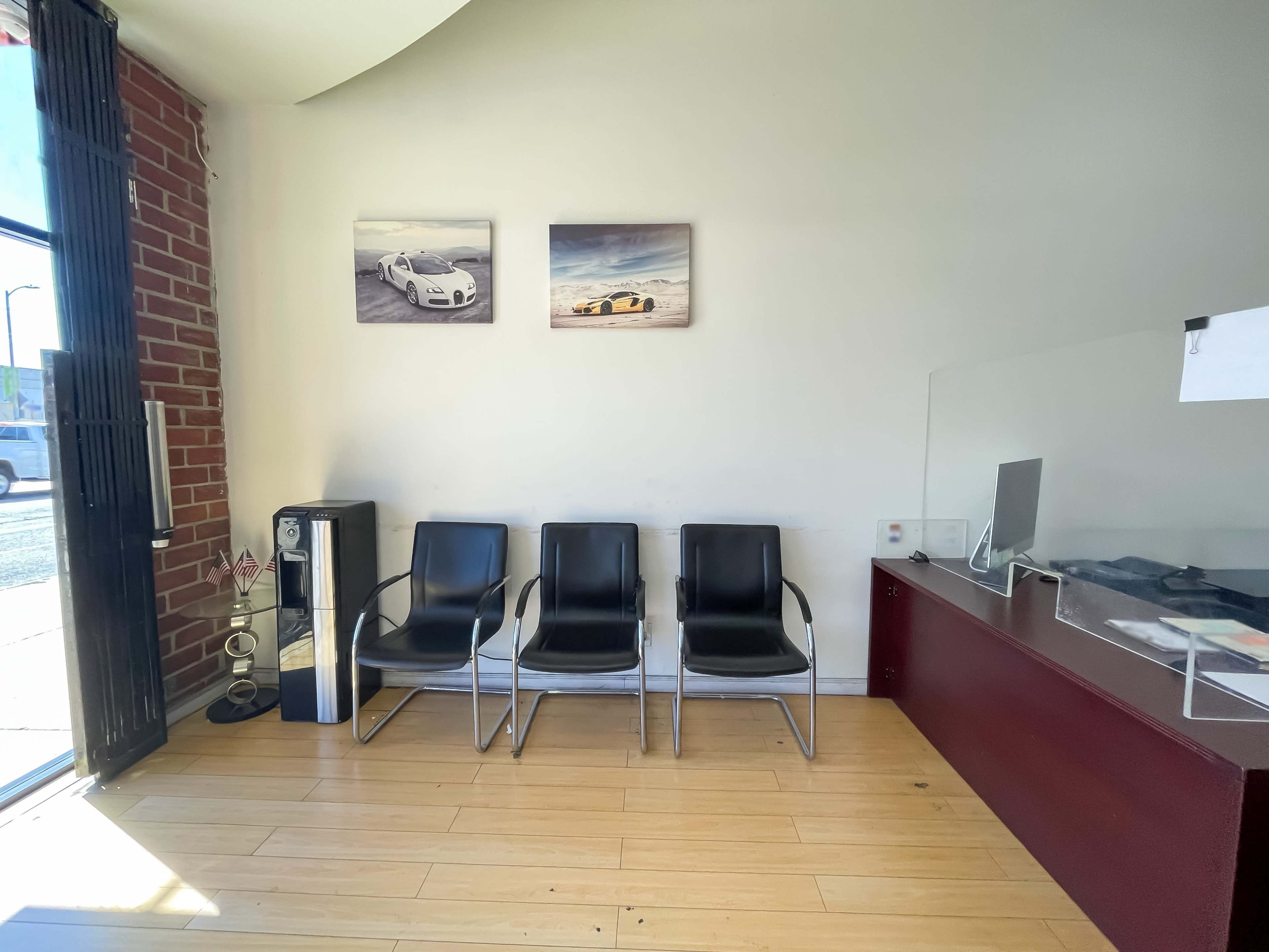 The image shows a waiting area with three black chairs, a refrigerator, and two framed car photographs on the wall.