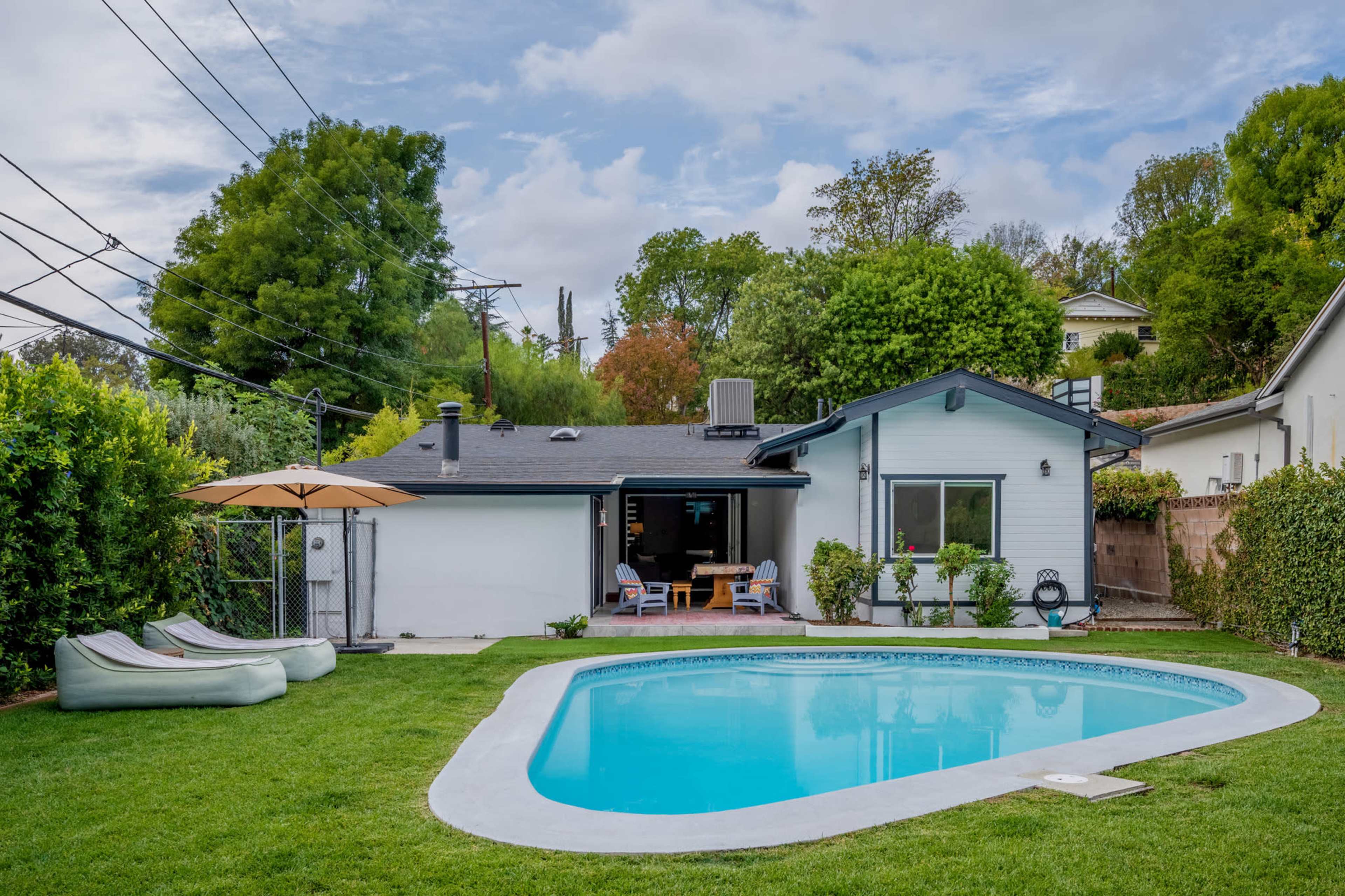 A backyard features a swimming pool surrounded by green grass, lounge chairs, and a patio area with a table and umbrella.