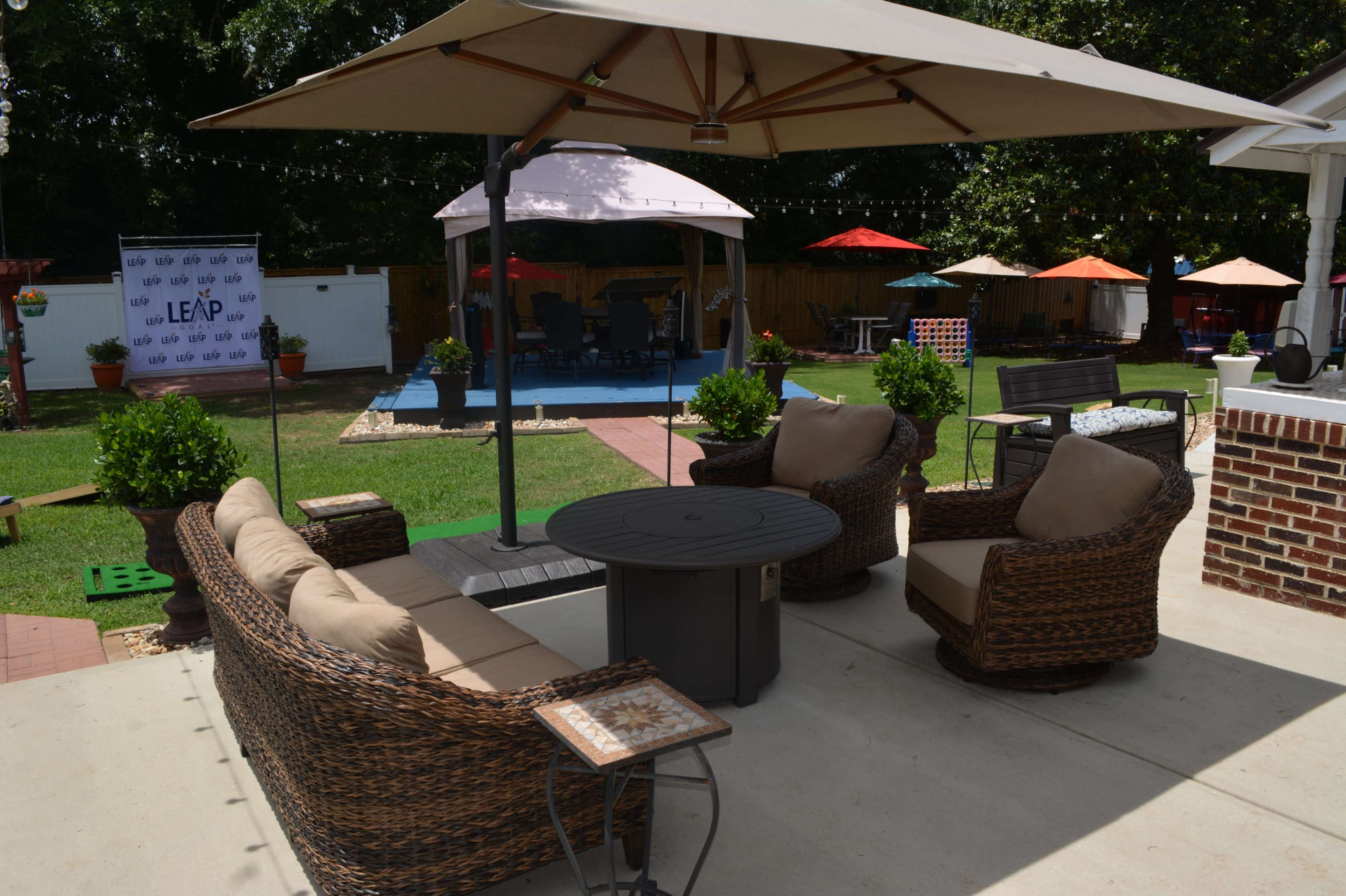 The image shows a backyard patio area with seating arrangements, an umbrella, and various types of outdoor furniture in a landscaped yard.