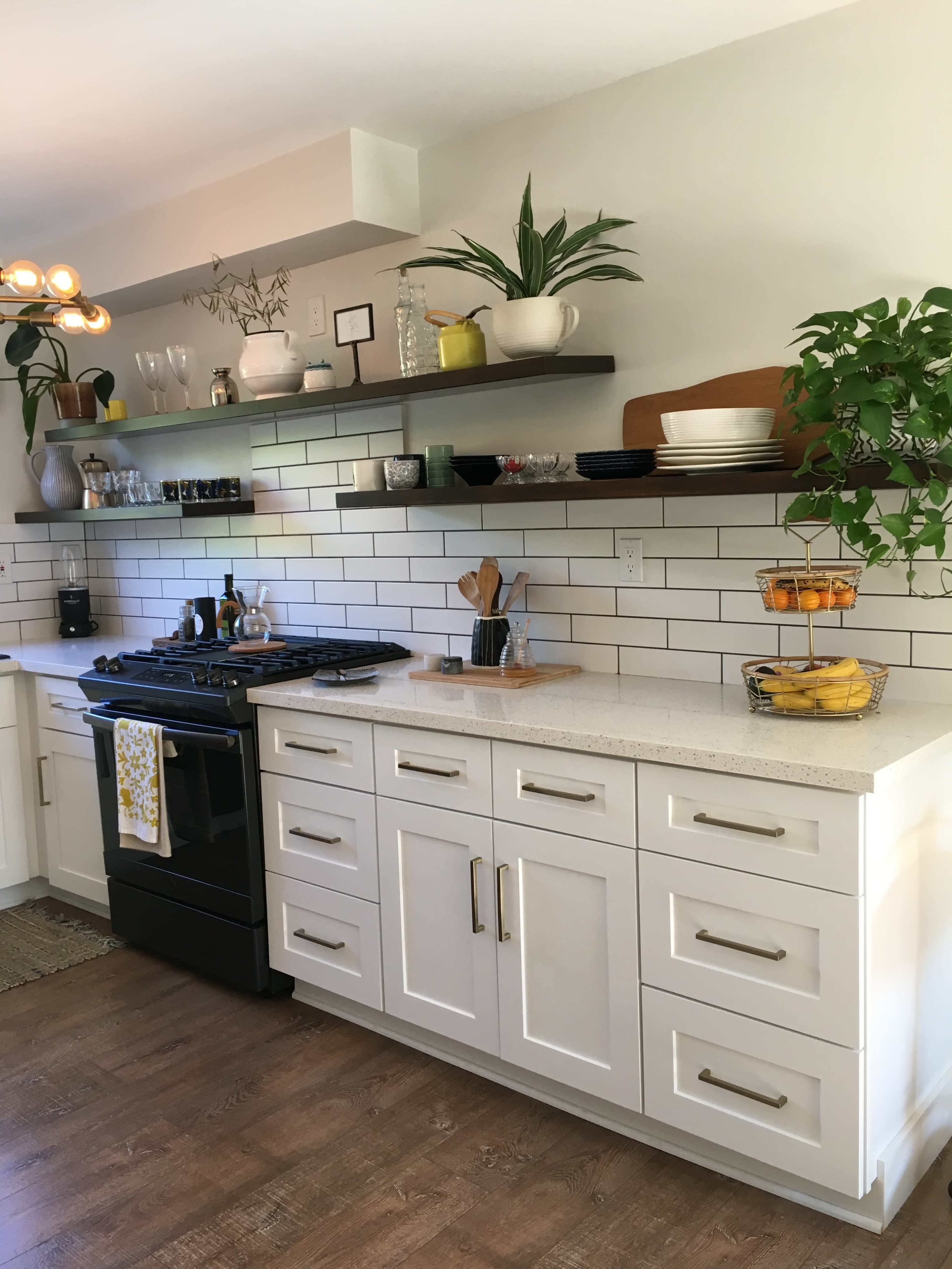 A modern kitchen features a black stove, white cabinets, open shelving with dishes and plants, and a countertop with utensils and a bowl of fruit.