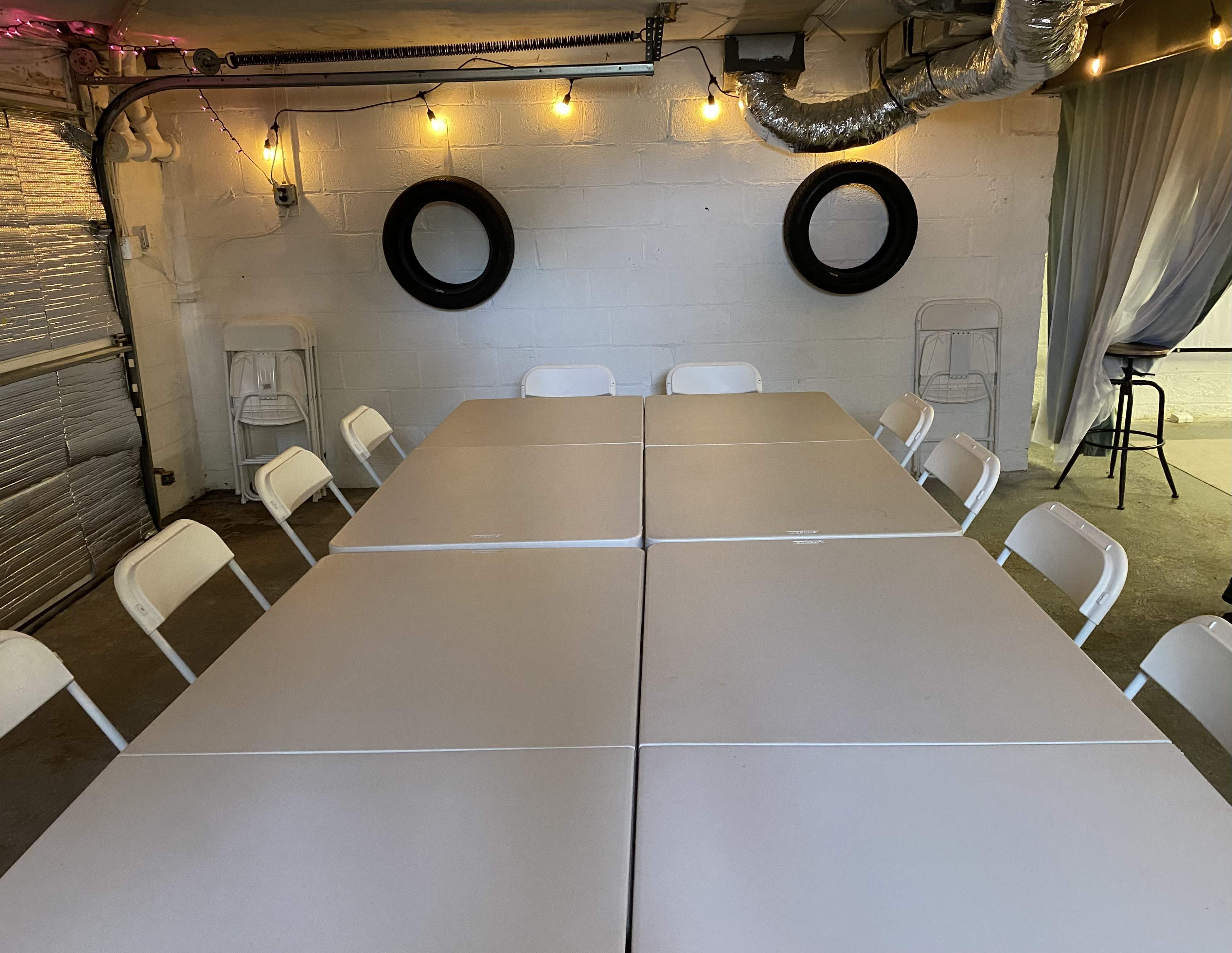 A set of white folding tables arranged in a rectangular formation under lights in a basement-like space with two tire-shaped wall decorations.