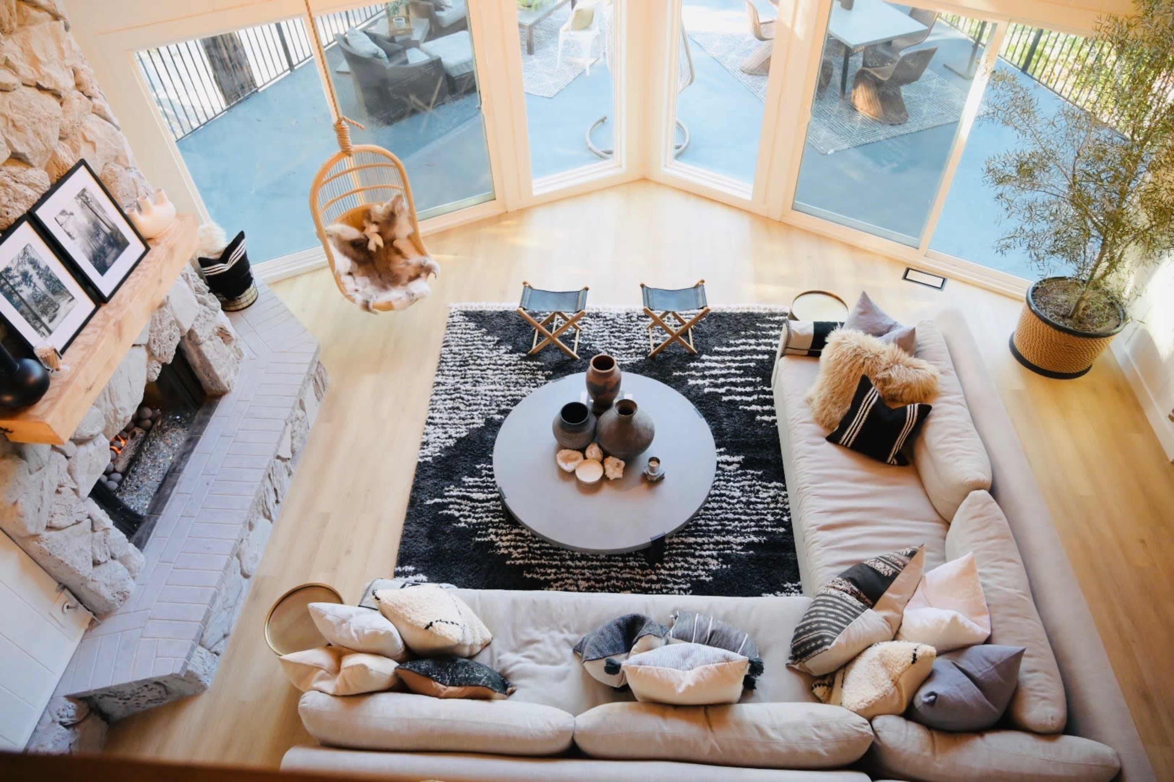 A spacious living room features a large sectional sofa, a round coffee table, and a stone fireplace, all arranged around a black and white area rug, with large windows allowing ample natural light.