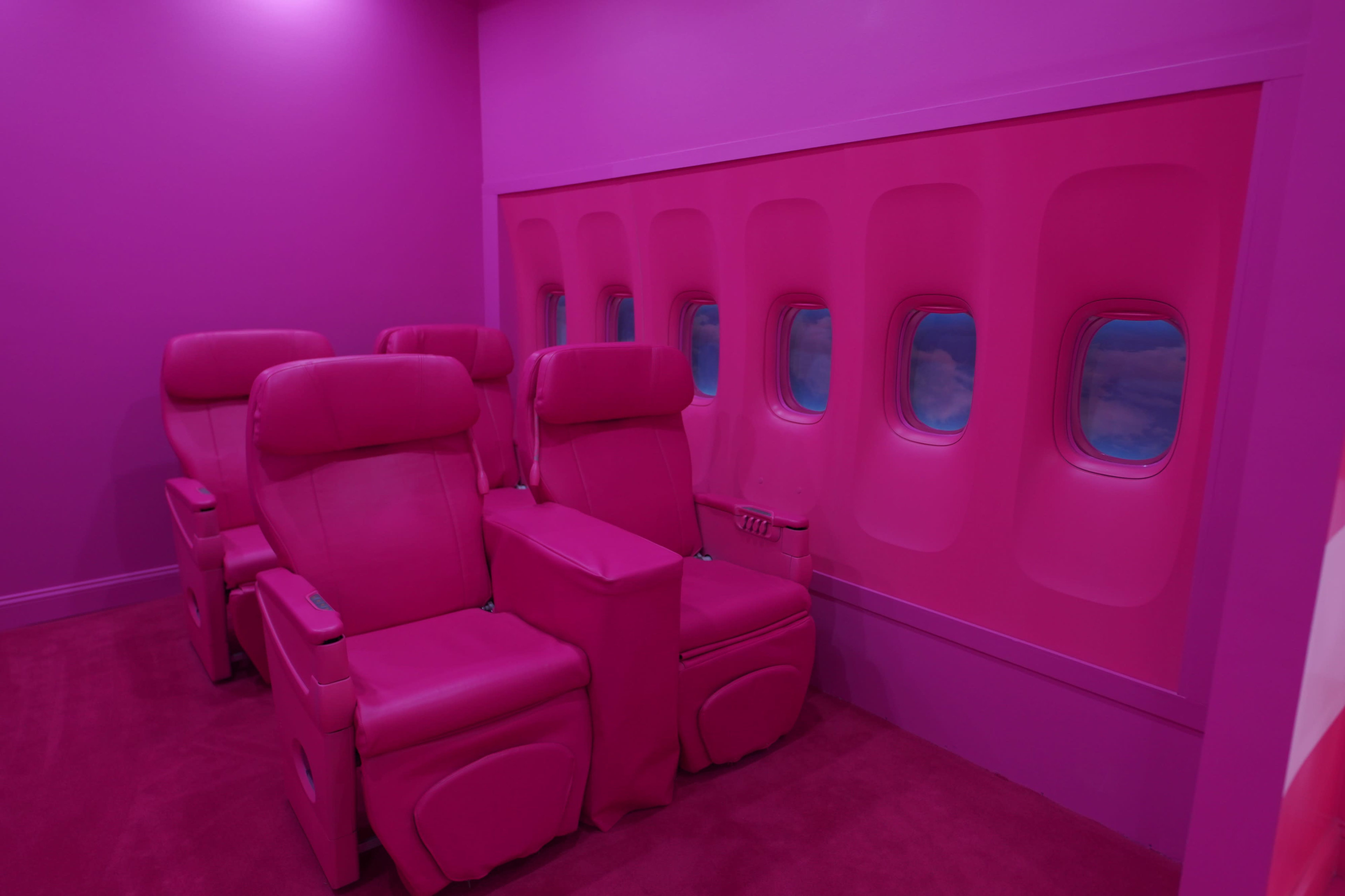 A small room with pink walls and four pink reclining airline seats arranged next to a wall featuring window-like cutouts displaying a blue sky.