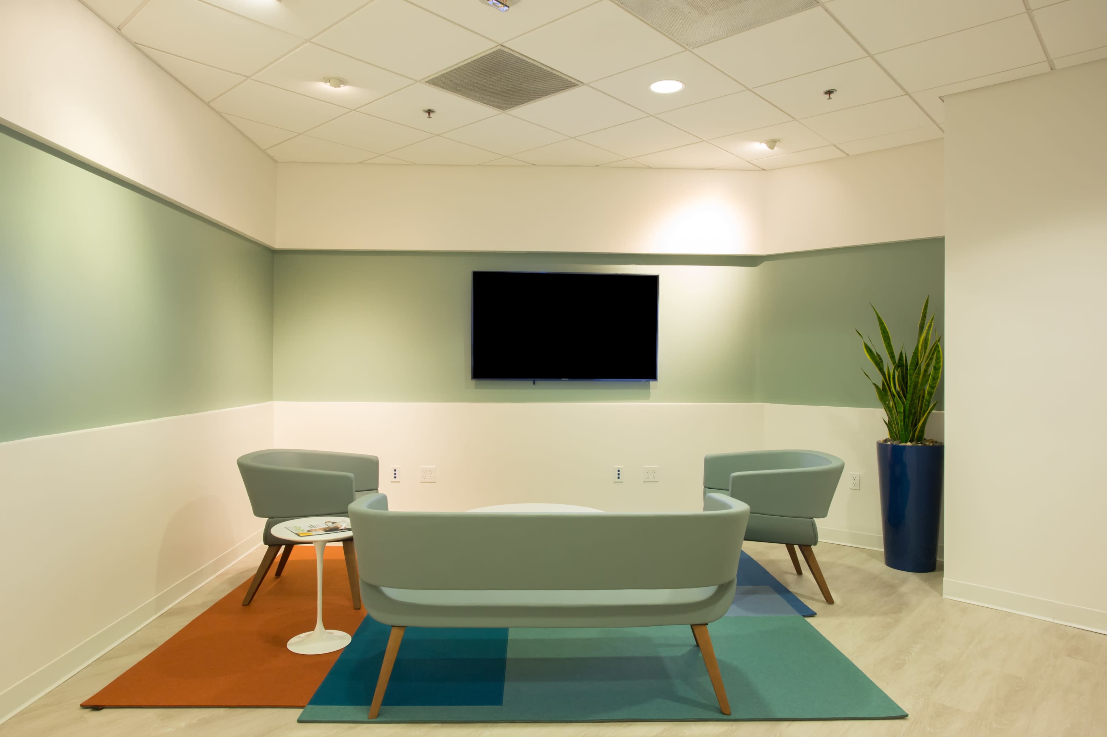 A modern, minimalist waiting area with two light green chairs, a small round table, a blue plant pot, and a wall-mounted television.