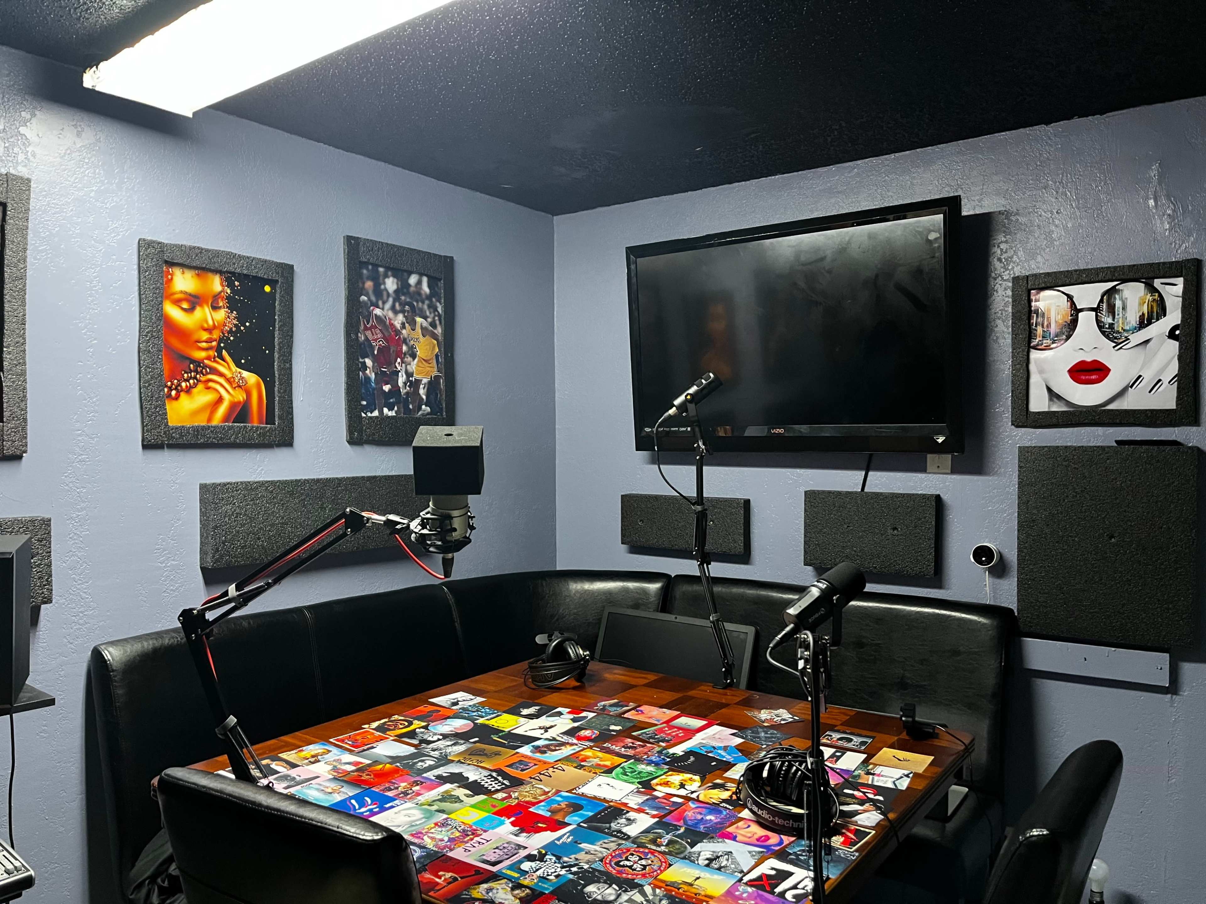 The image shows a recording studio with a table covered in album covers, microphones on stands, and art displayed on the walls.