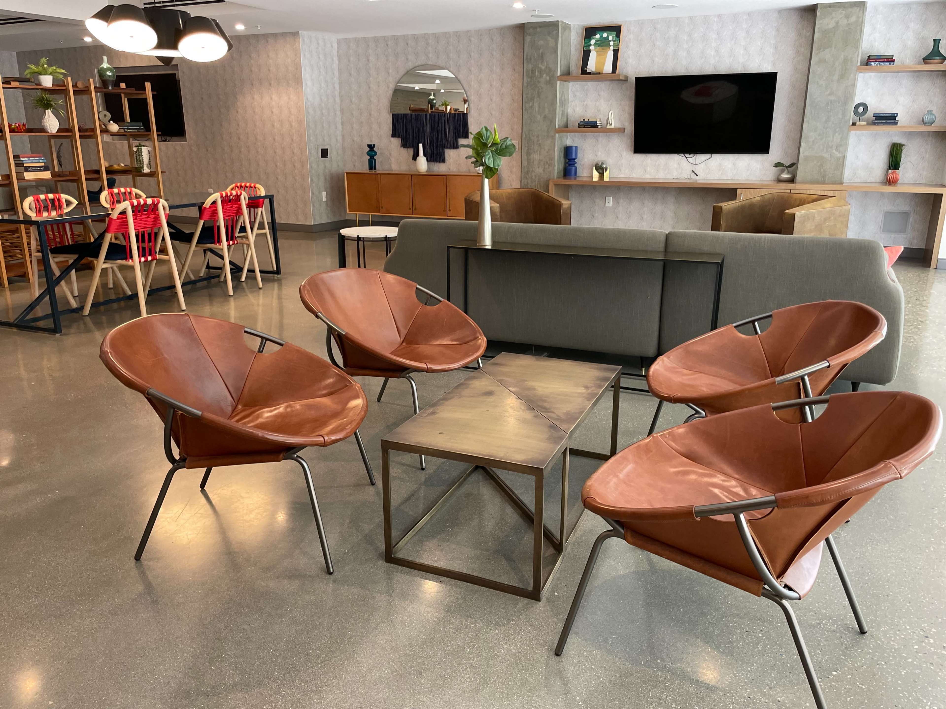 A modern lounge area features four brown leather chairs arranged around a square coffee table, with a backdrop of stylish furniture and decor.