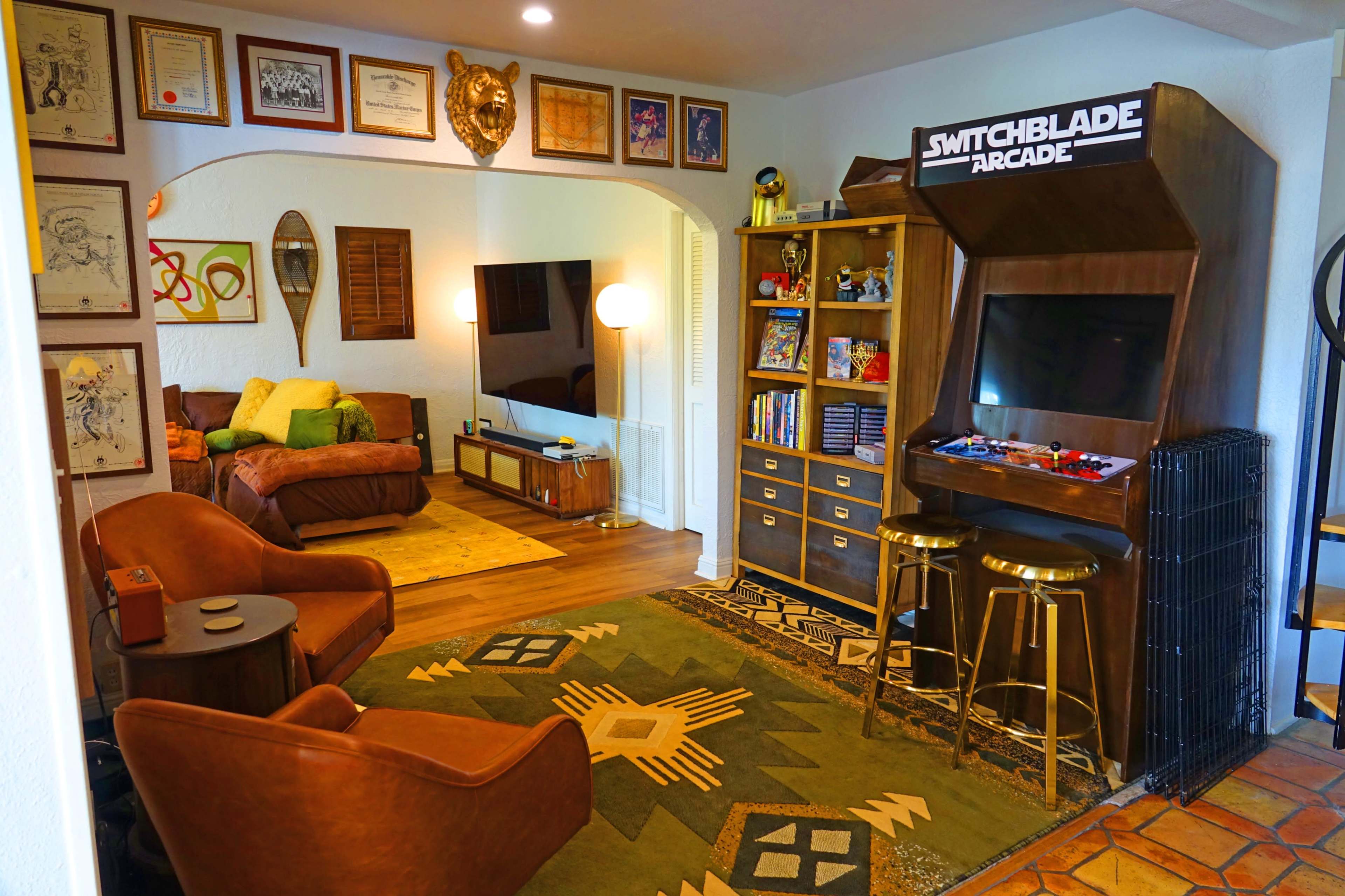 The image showcases a cozy living room with a retro arcade cabinet, a comfortable seating area, and framed artwork on the walls.