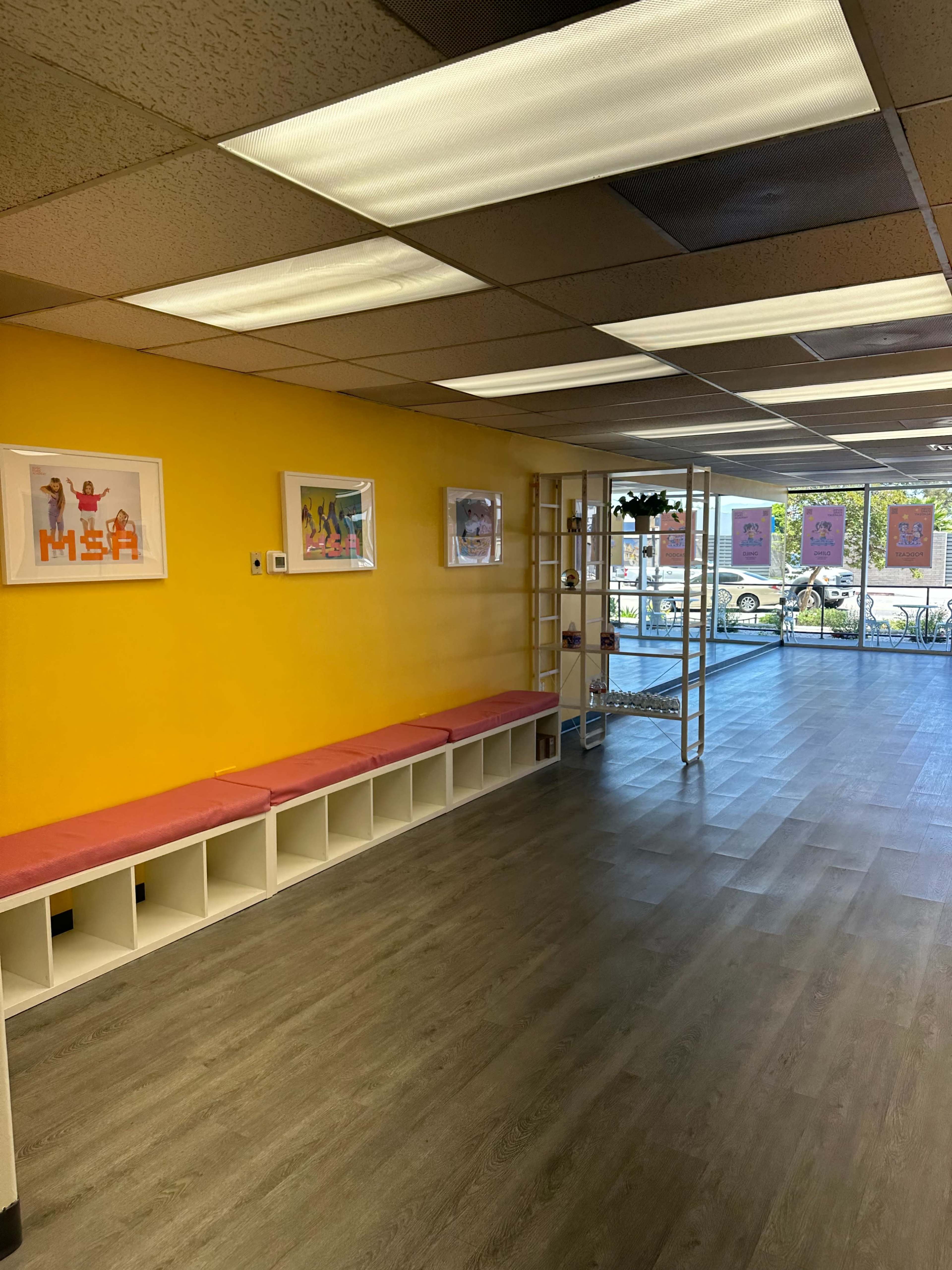 The image shows a brightly colored yellow wall in a room with low benches, framed artwork, and large windows allowing natural light to enter.