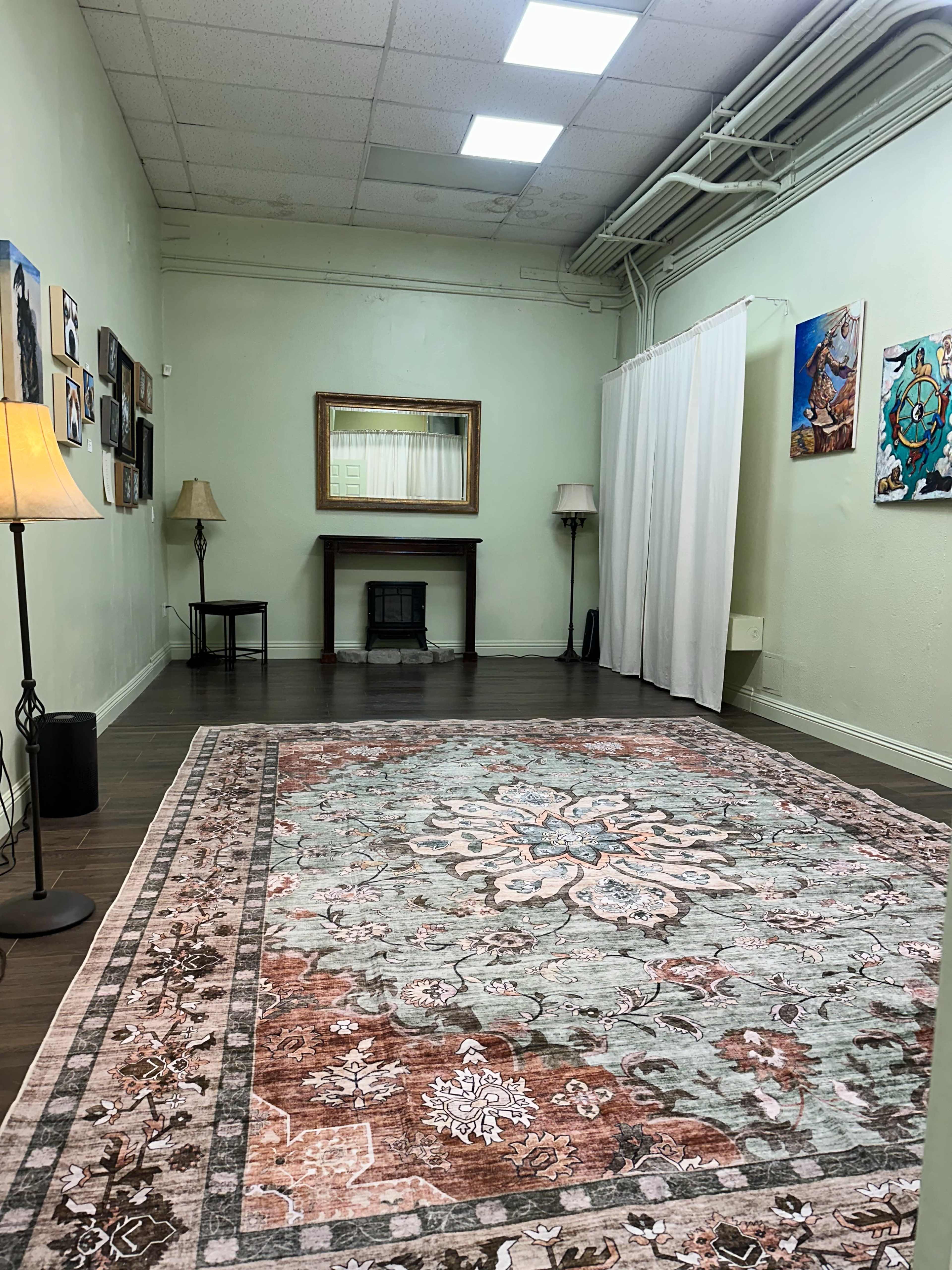 The image shows a spacious room with light green walls, a large patterned rug on the floor, a mirror above a small fireplace, and various artworks displayed on the walls.
