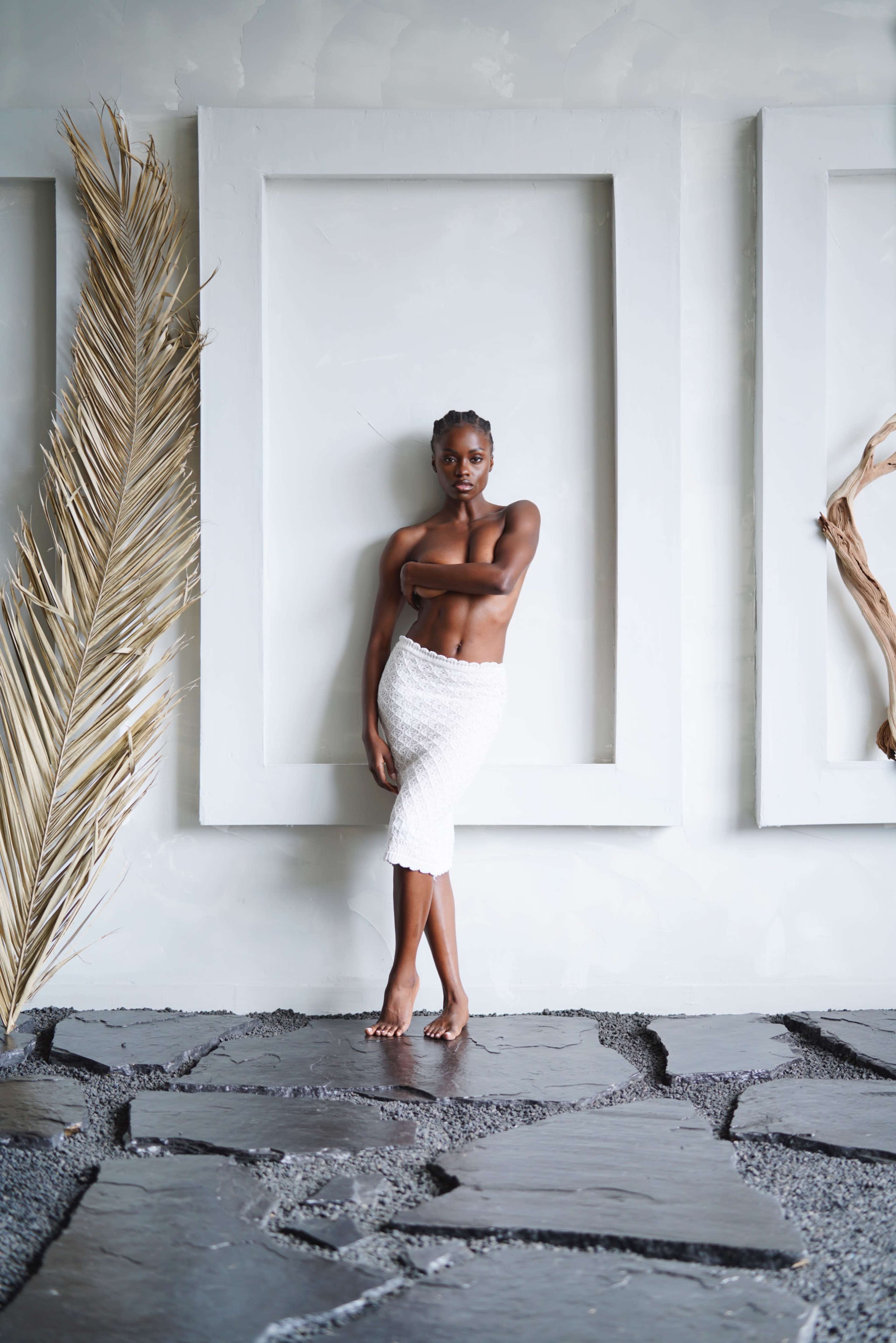 A woman stands barefoot against a textured wall with large, white frames and natural elements nearby.