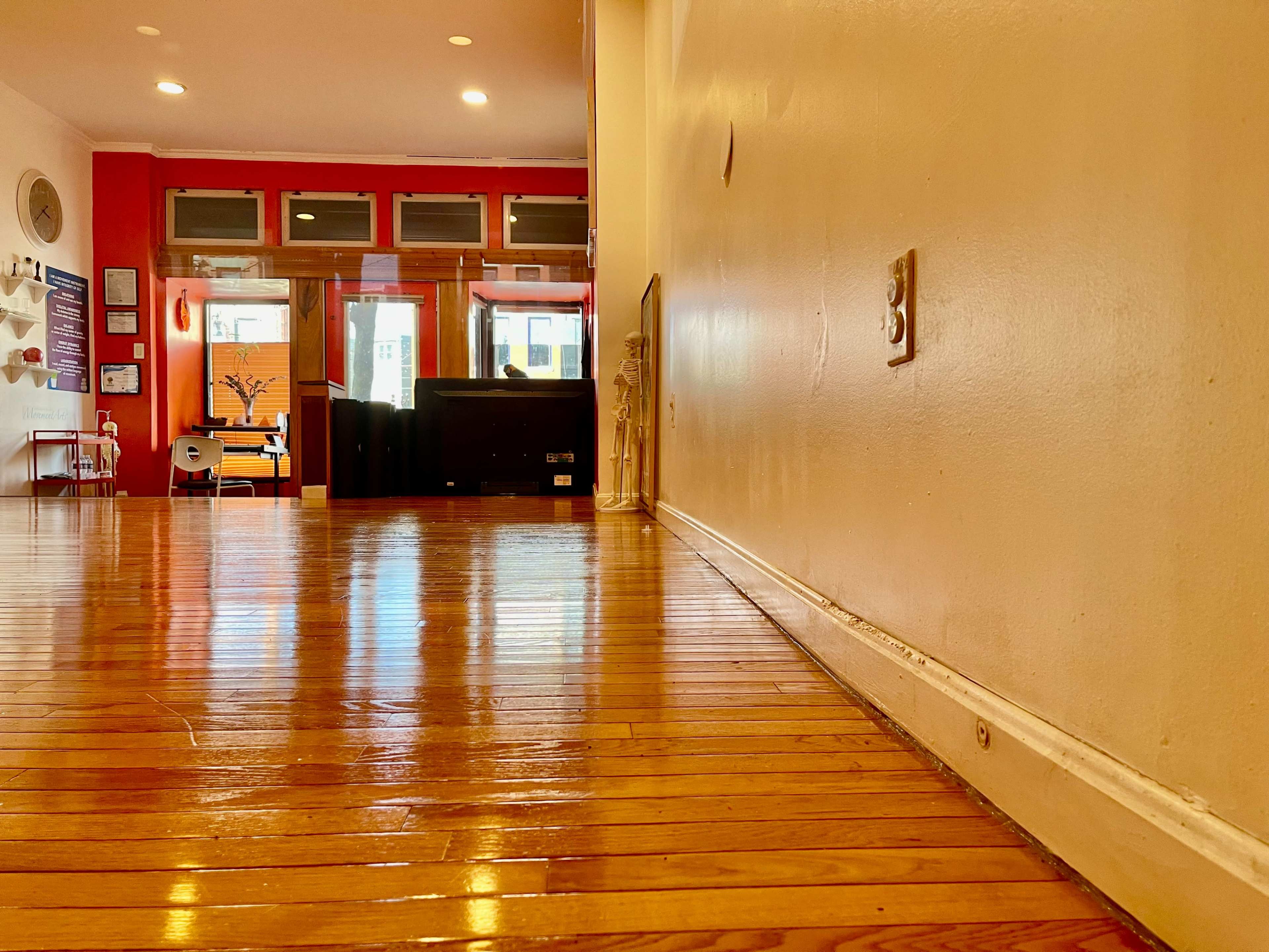 The image shows a polished wooden floor with a low angle view of a wall leading to an open area featuring windows and furniture in the background.