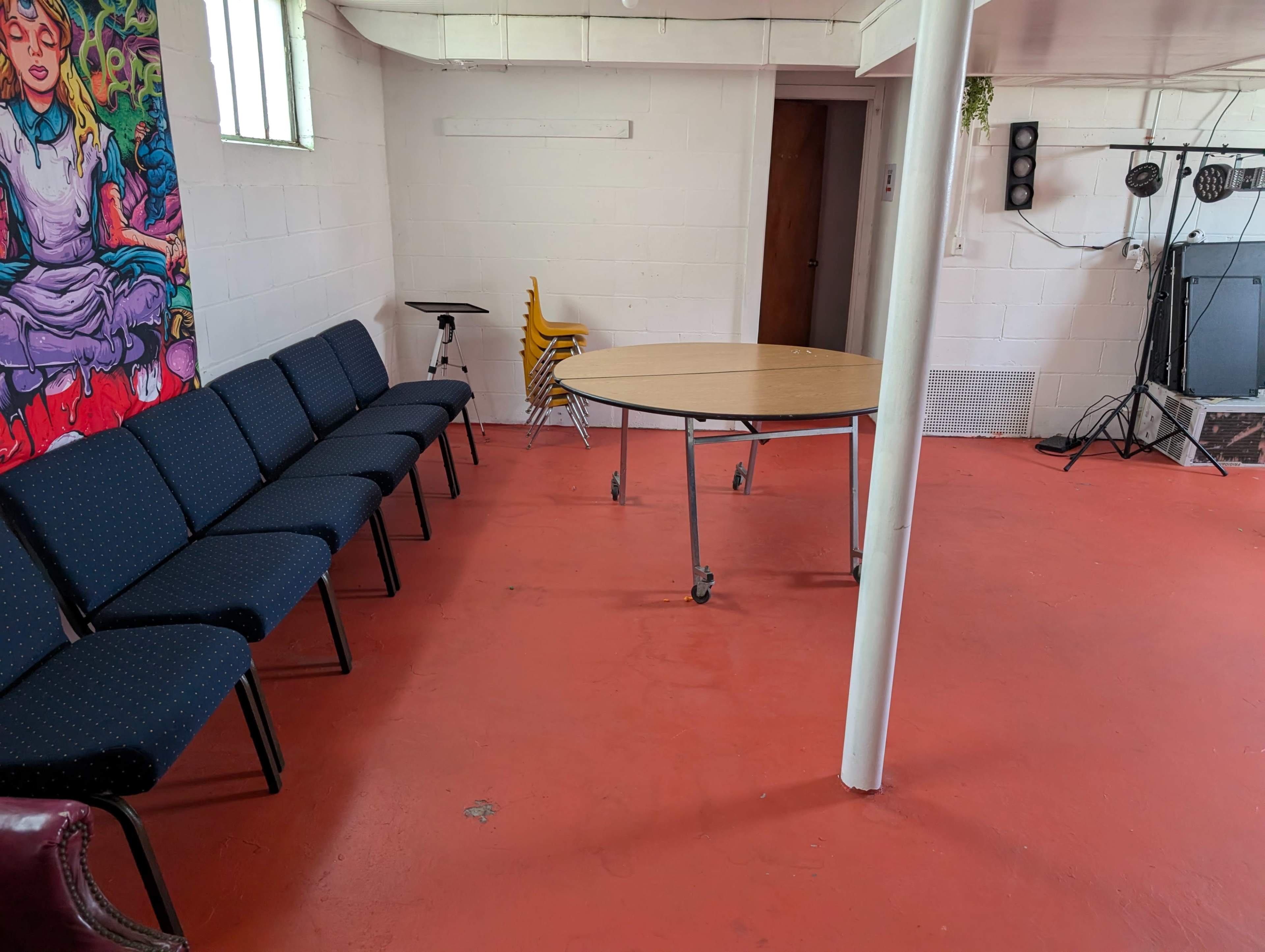 The image shows a room with a round table on wheels in the center, surrounded by blue chairs, and a vibrant mural on the wall.