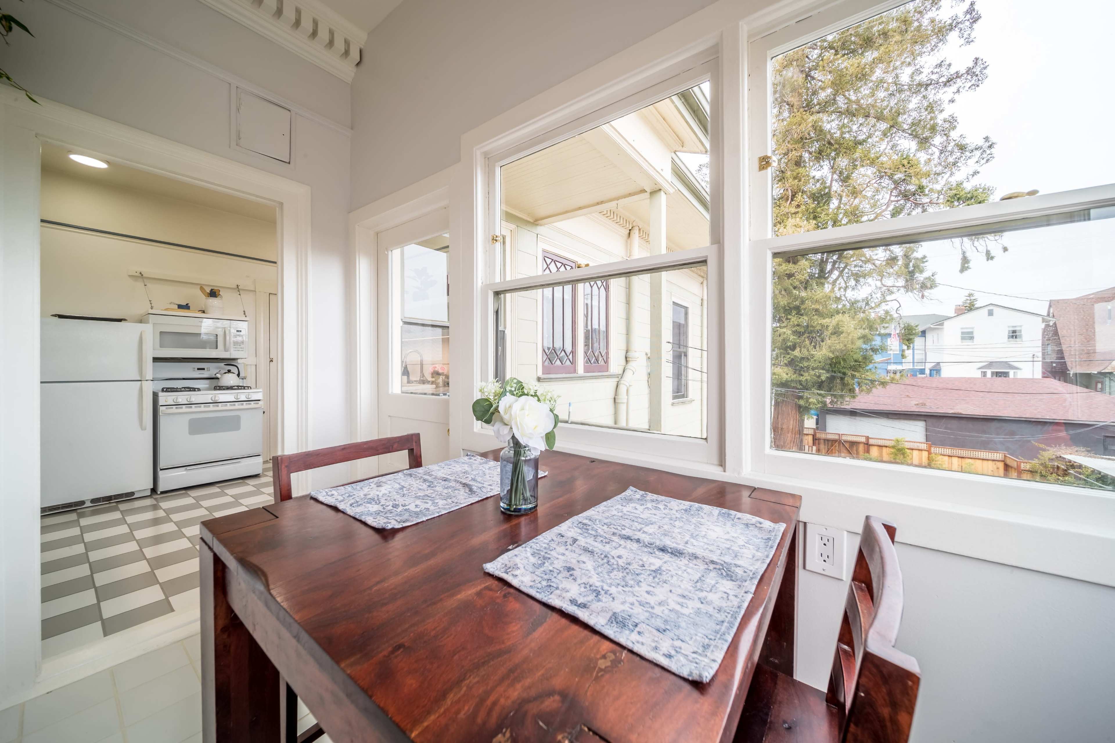 A wooden dining table with placemats is positioned near large windows, overlooking a kitchen area that includes white appliances and checkered flooring.