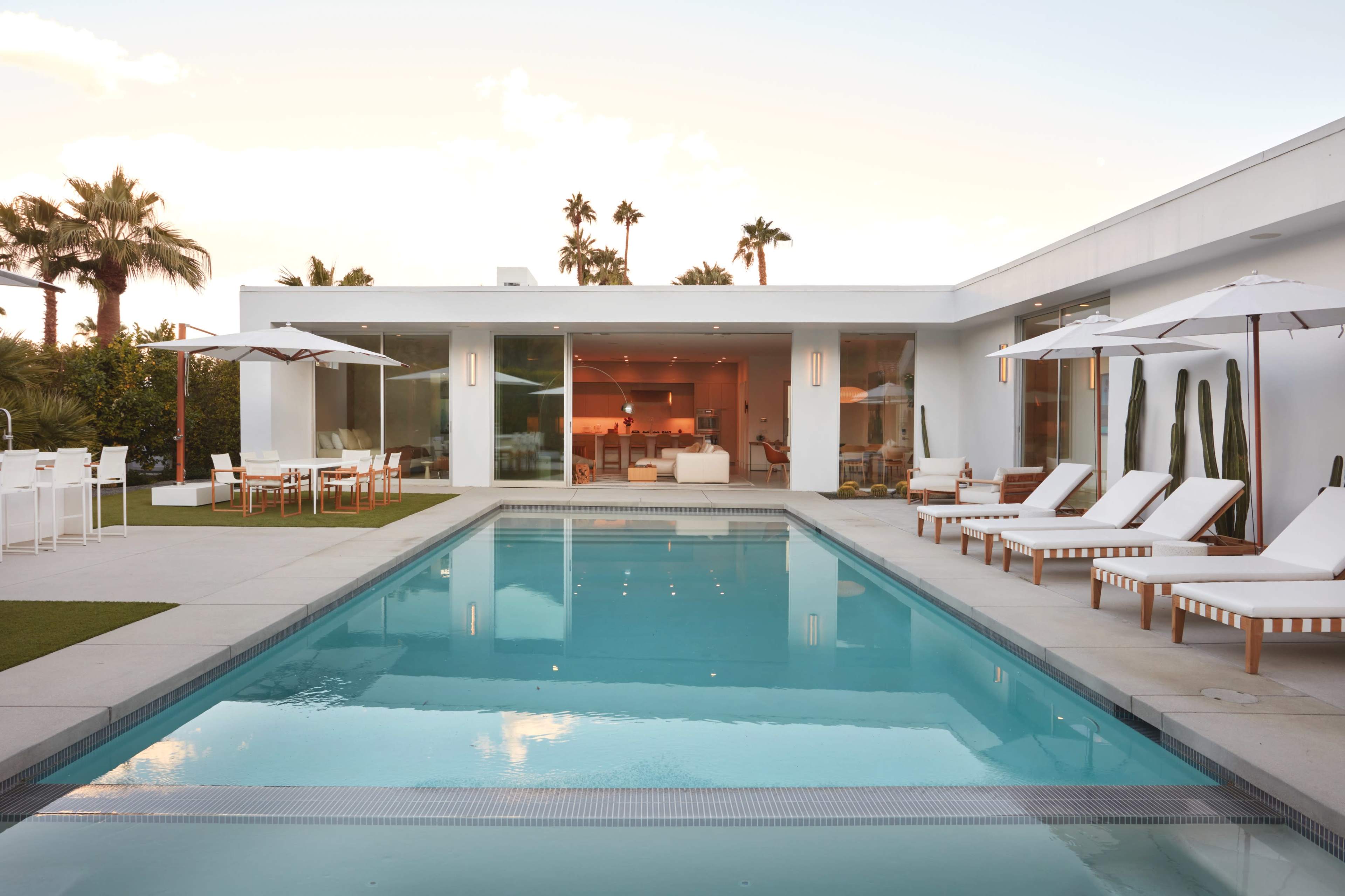 The image shows a modern home with a swimming pool, surrounded by lounge chairs and umbrellas, set against a backdrop of palm trees.