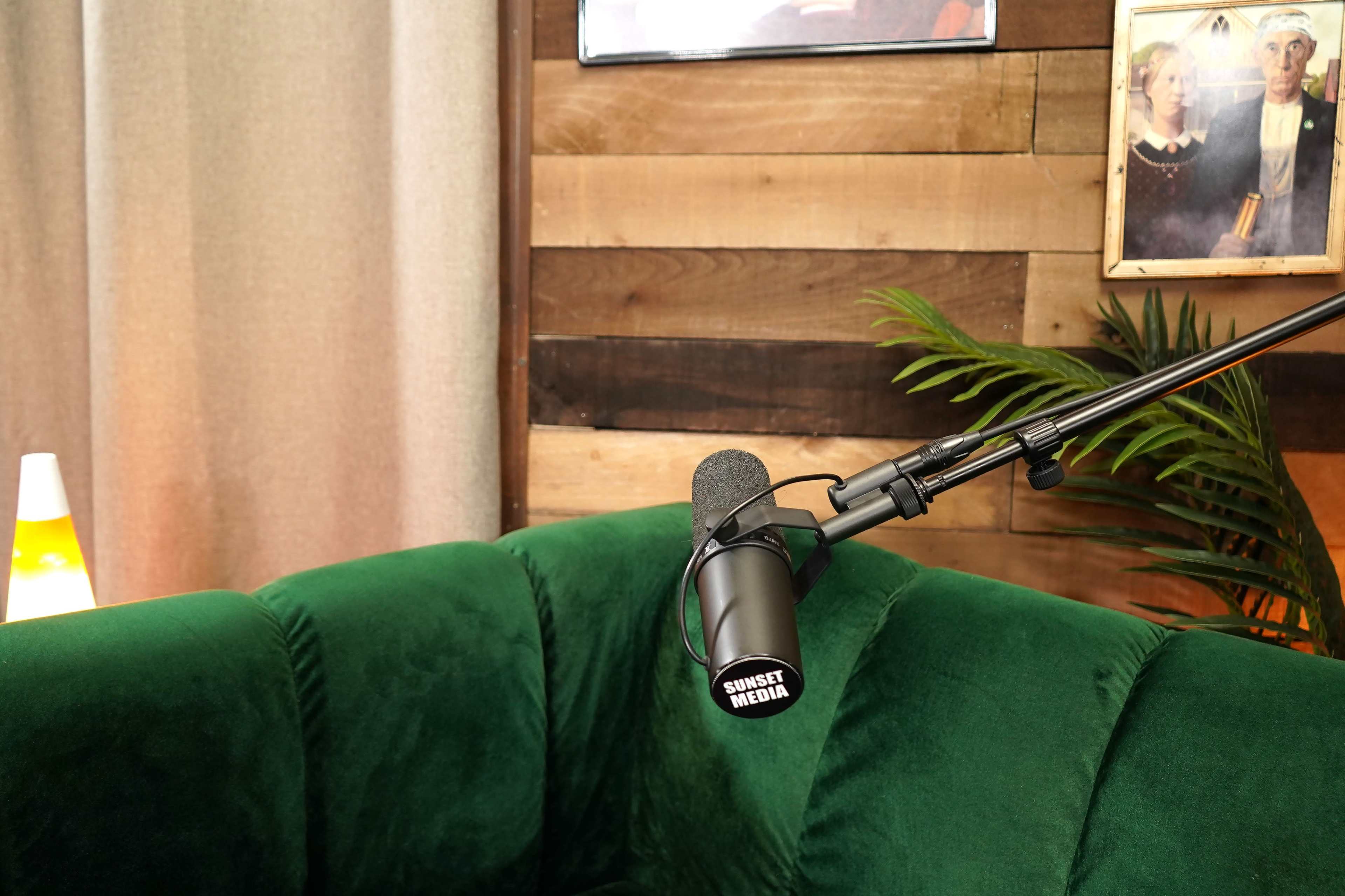 A green velvet couch sits against a wooden wall with a microphone labeled "SUNSET MEDIA" positioned nearby, complemented by decorative elements like a plant and framed pictures.