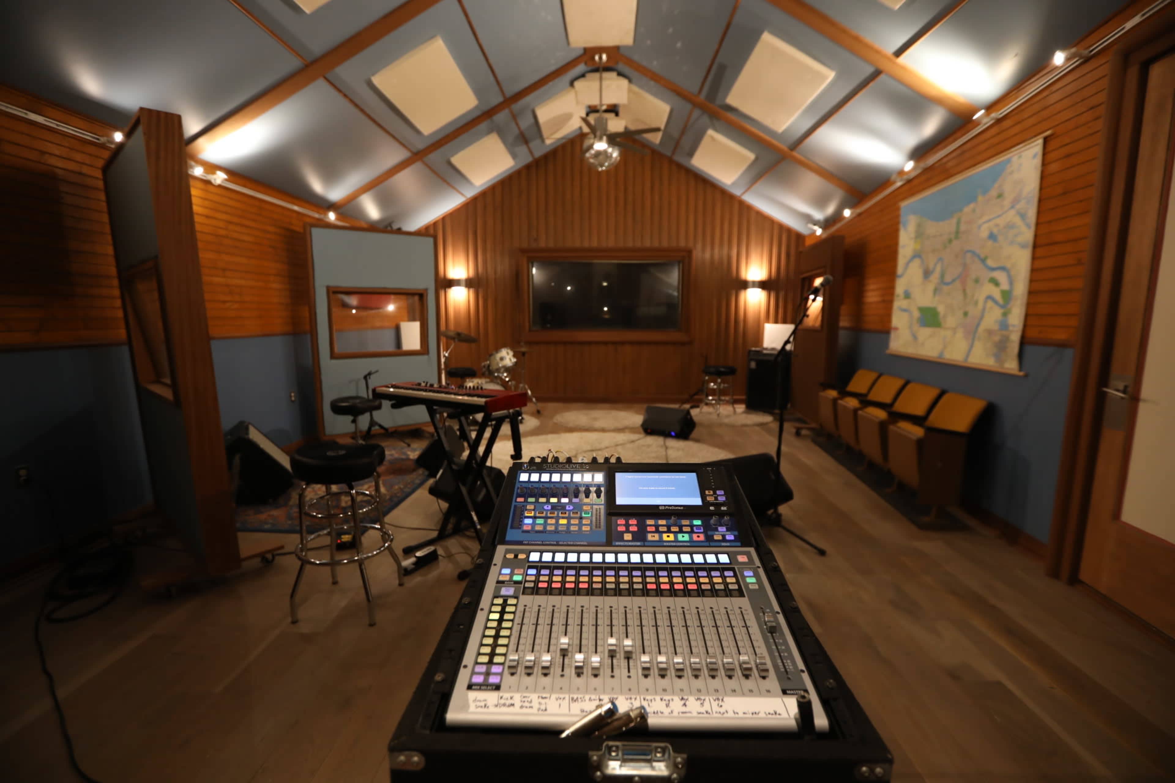 The image shows the interior of a recording studio featuring a digital mixing console in the foreground, with musical instruments and seating arranged in the background.