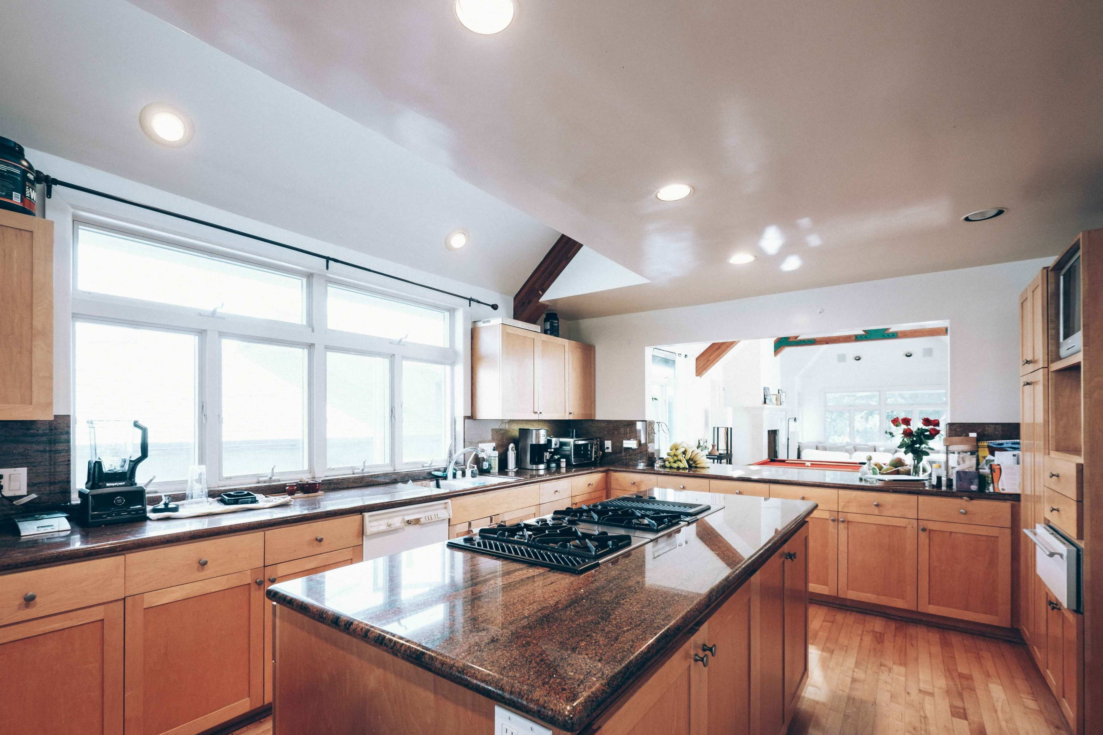 A spacious kitchen with wooden cabinets, a central island with a gas stove, and large windows allowing natural light to enter.