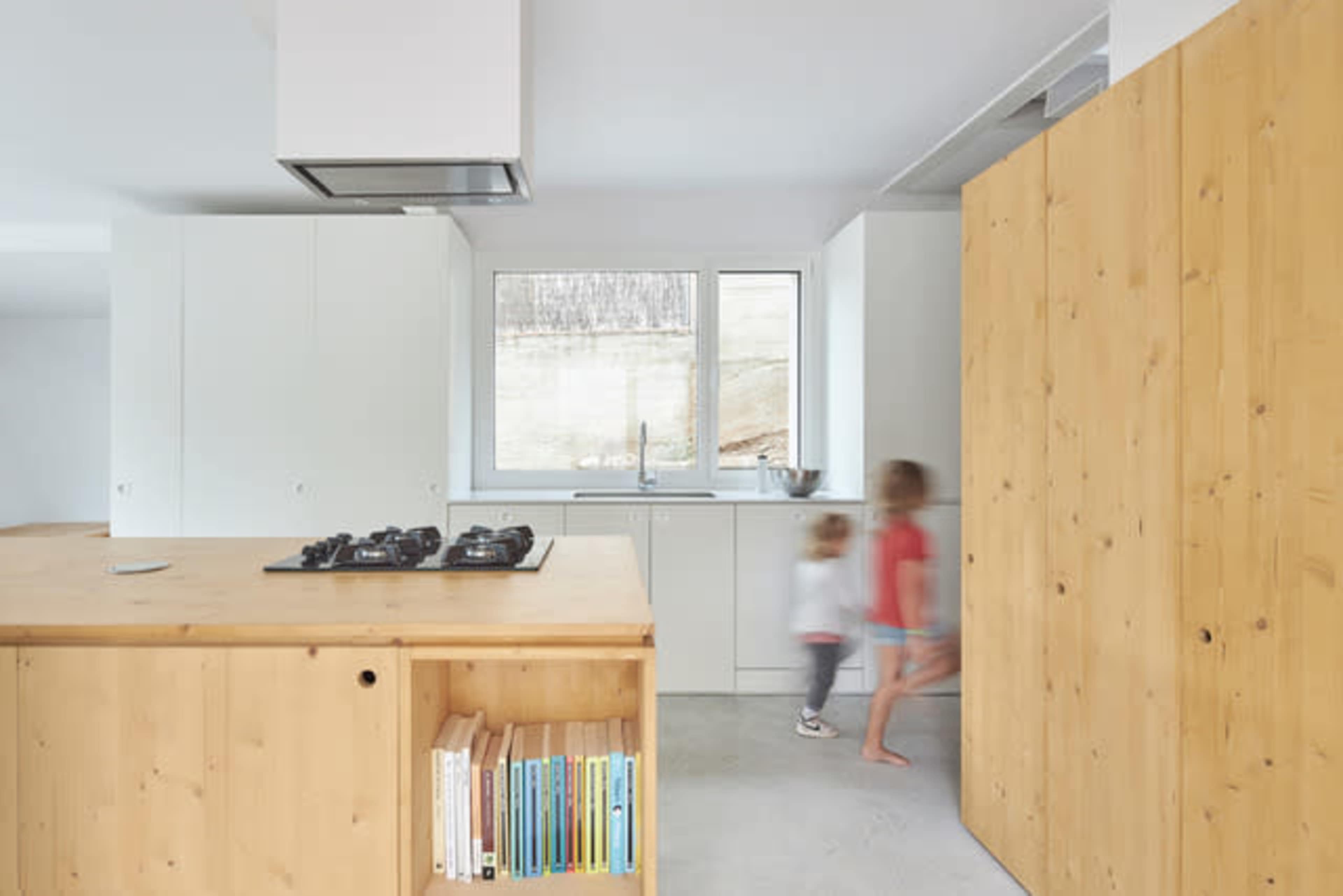 A modern kitchen features a wooden island with a gas stove, white cabinetry, and two children walking past a large window.
