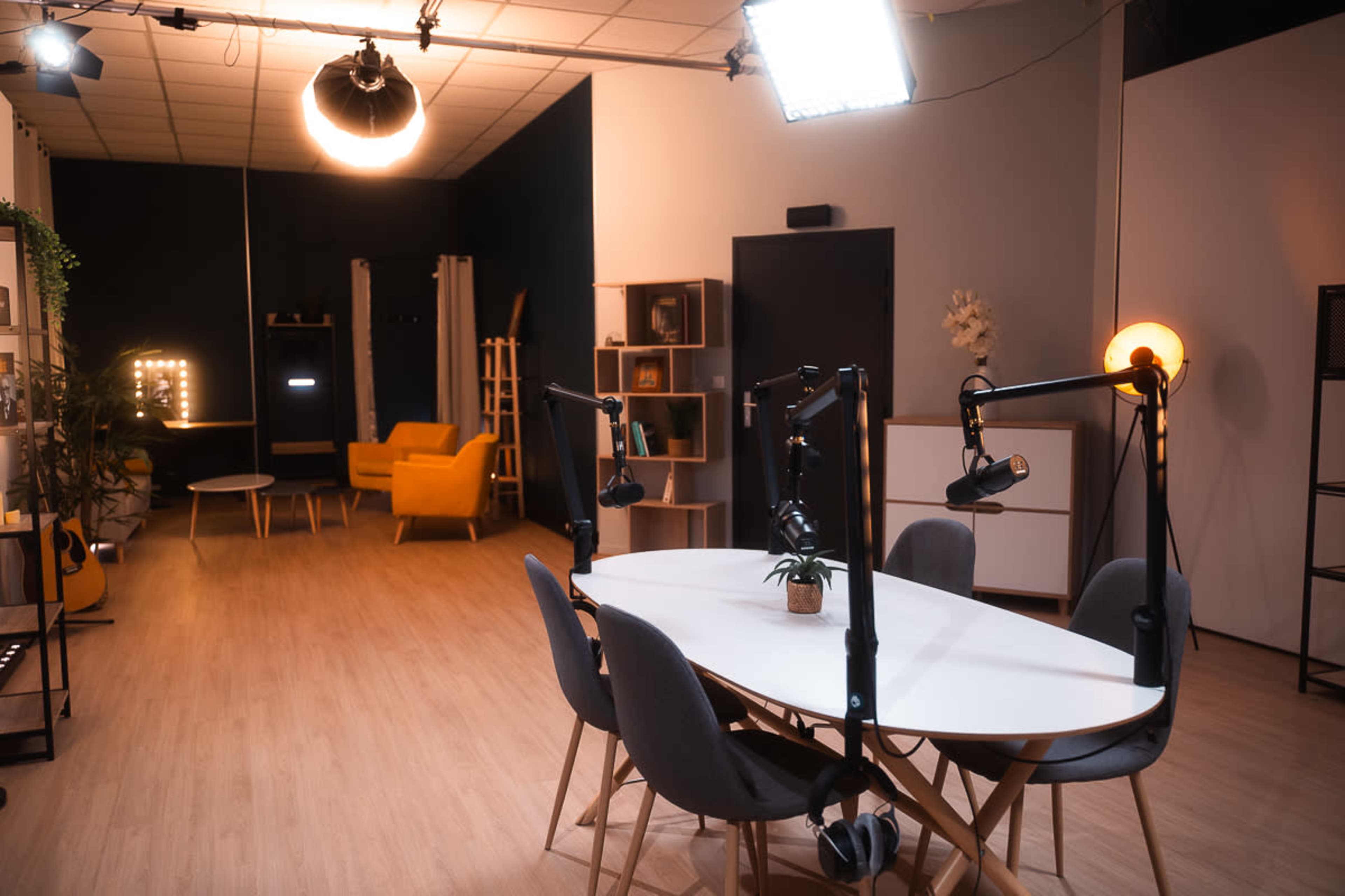 A modern studio space features a large oval table surrounded by four chairs, illuminated by overhead lights, with a yellow sofa and decorative elements in the background.