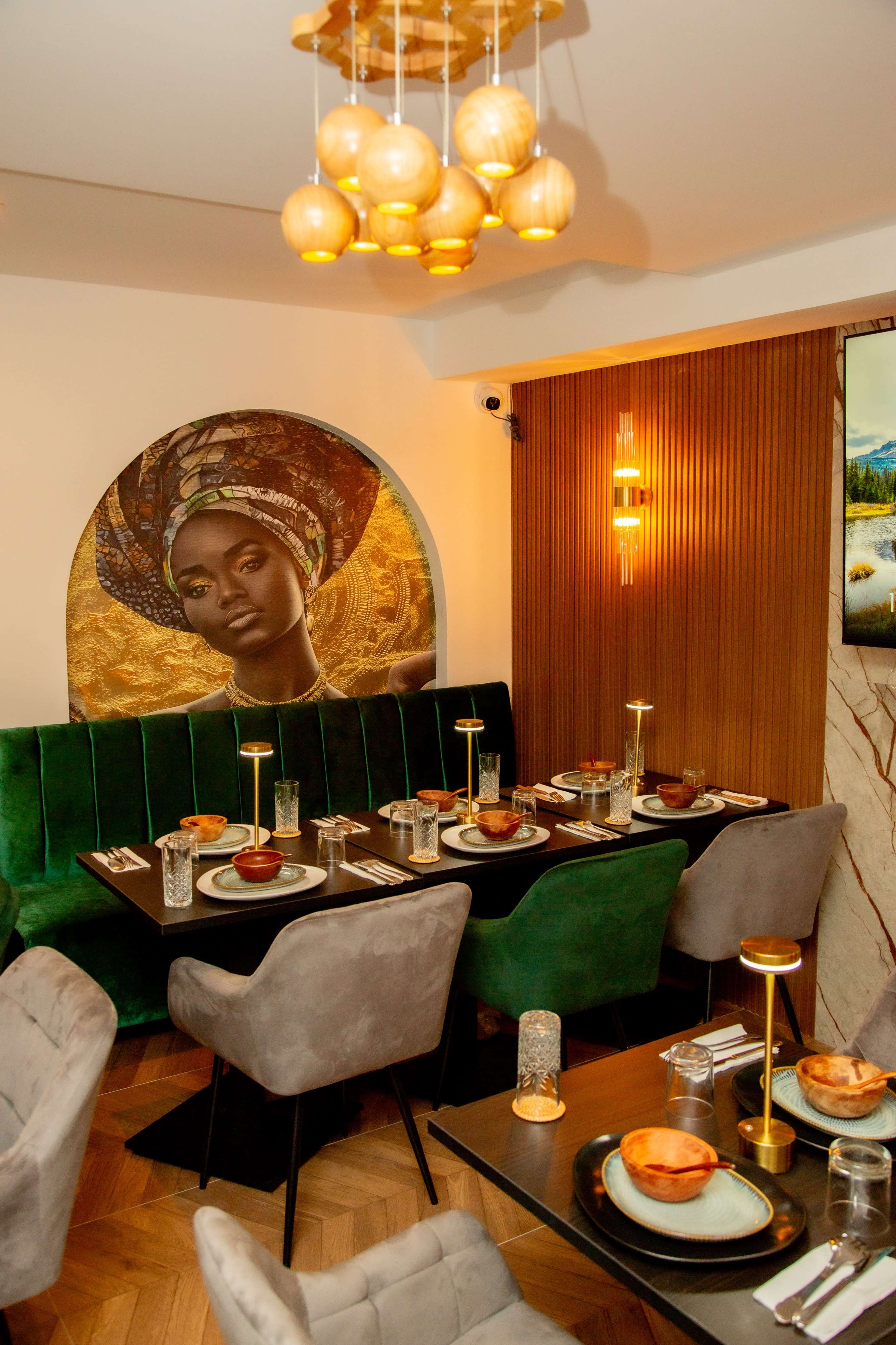 The image shows a restaurant interior featuring a green upholstered seating area, elegantly set tables with candles, and a large decorative wall piece of a woman with a headwrap.