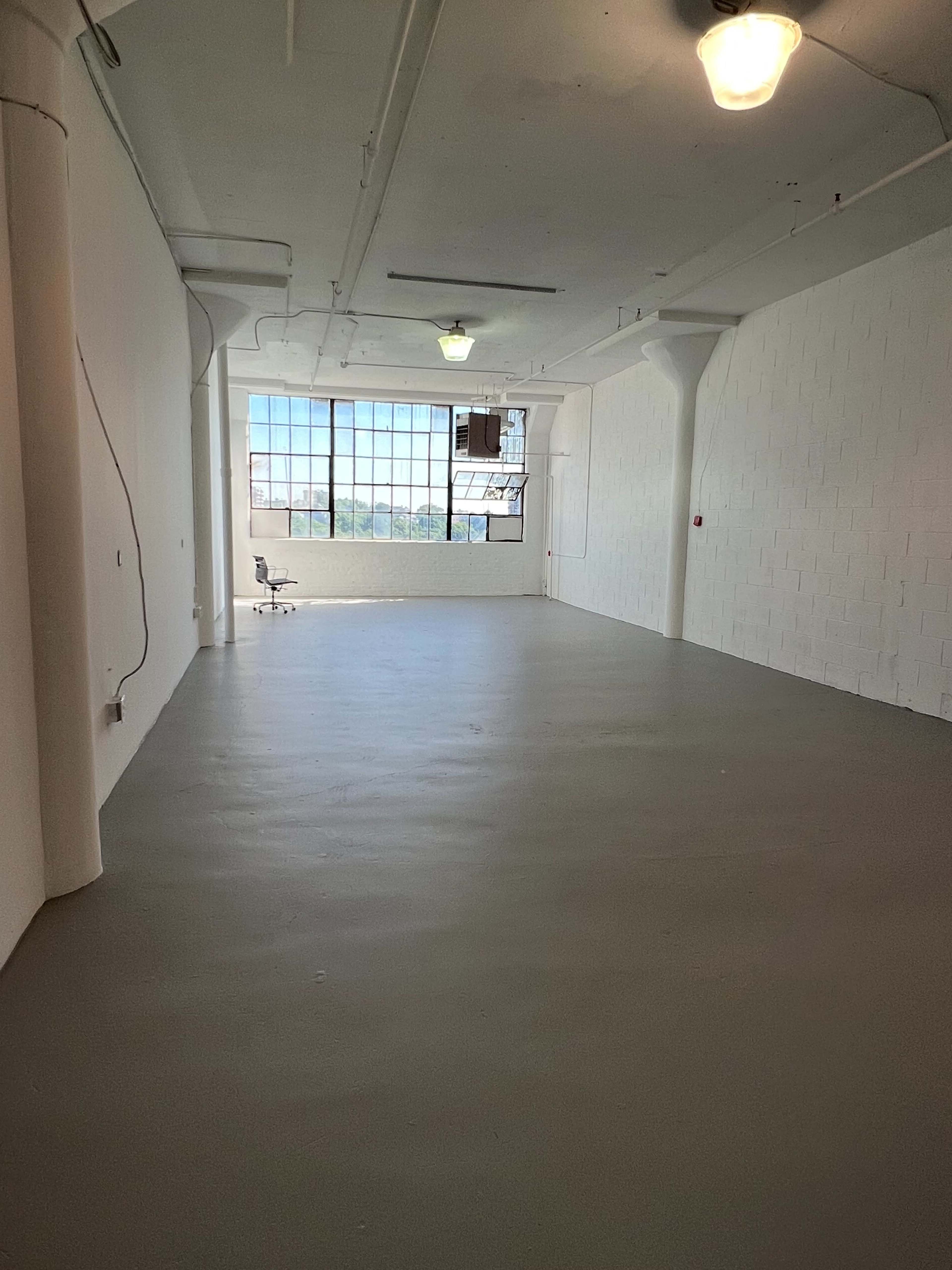 An empty industrial-style room features a large window and a single chair positioned in the corner.