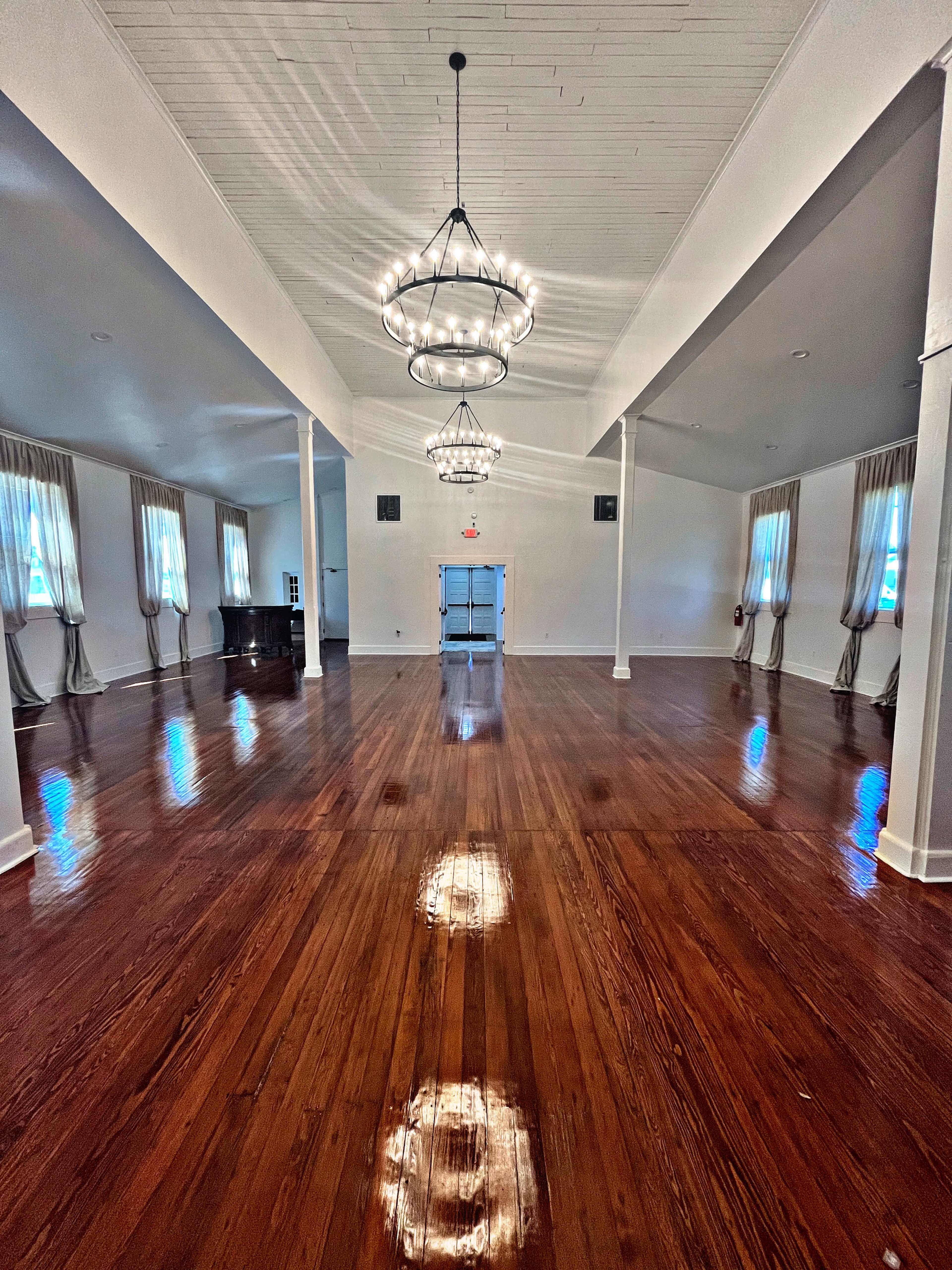 A spacious interior room with polished wooden floors, white walls, and chandelier lighting.