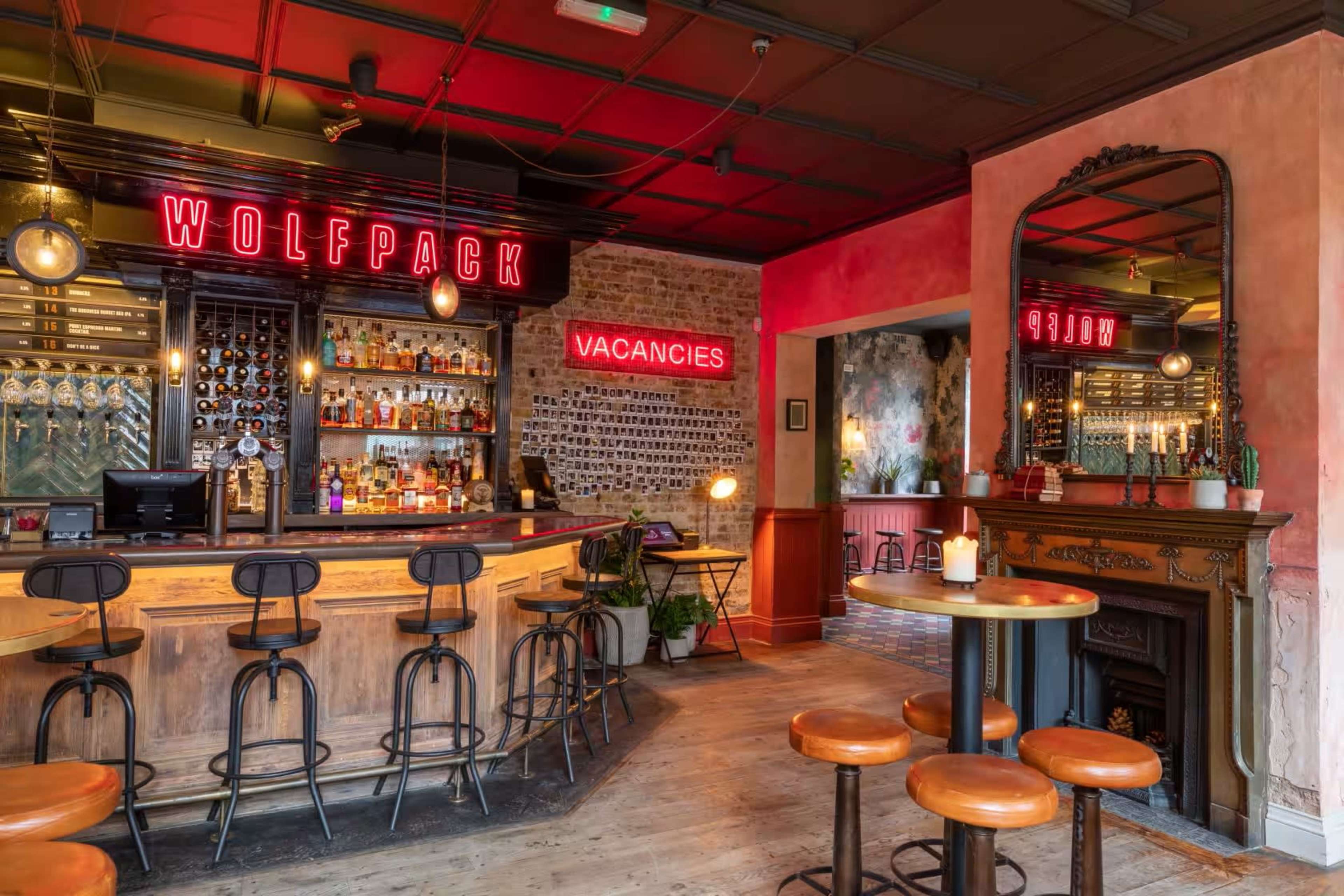 The image shows a bar area with a wooden counter, high stools, illuminated signage that reads "WOLFPACK" and "VACANCIES," and a mirror reflecting the interior decor.